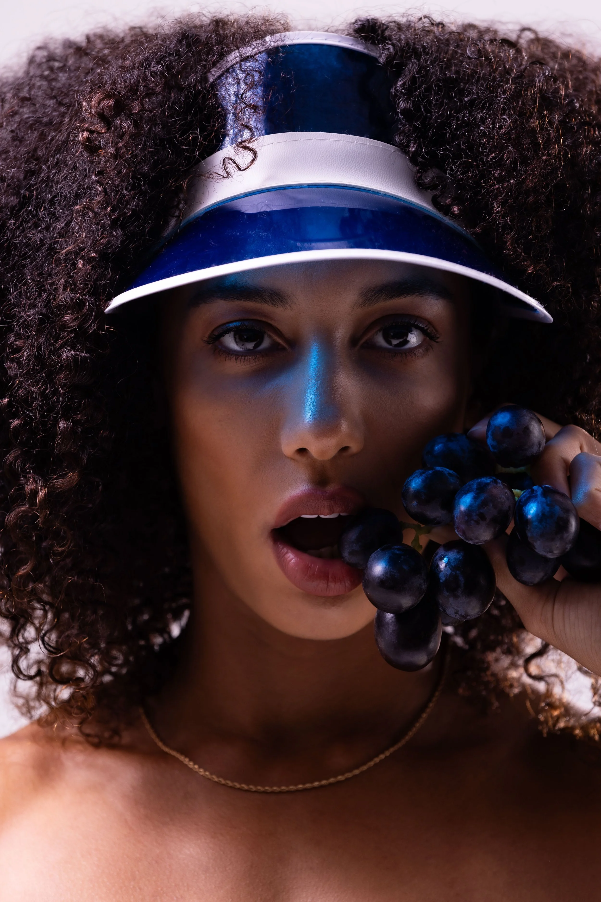 A woman with curly hair wearing a visor hat and holding a bunch of black grapes near her face, with her mouth slightly open.