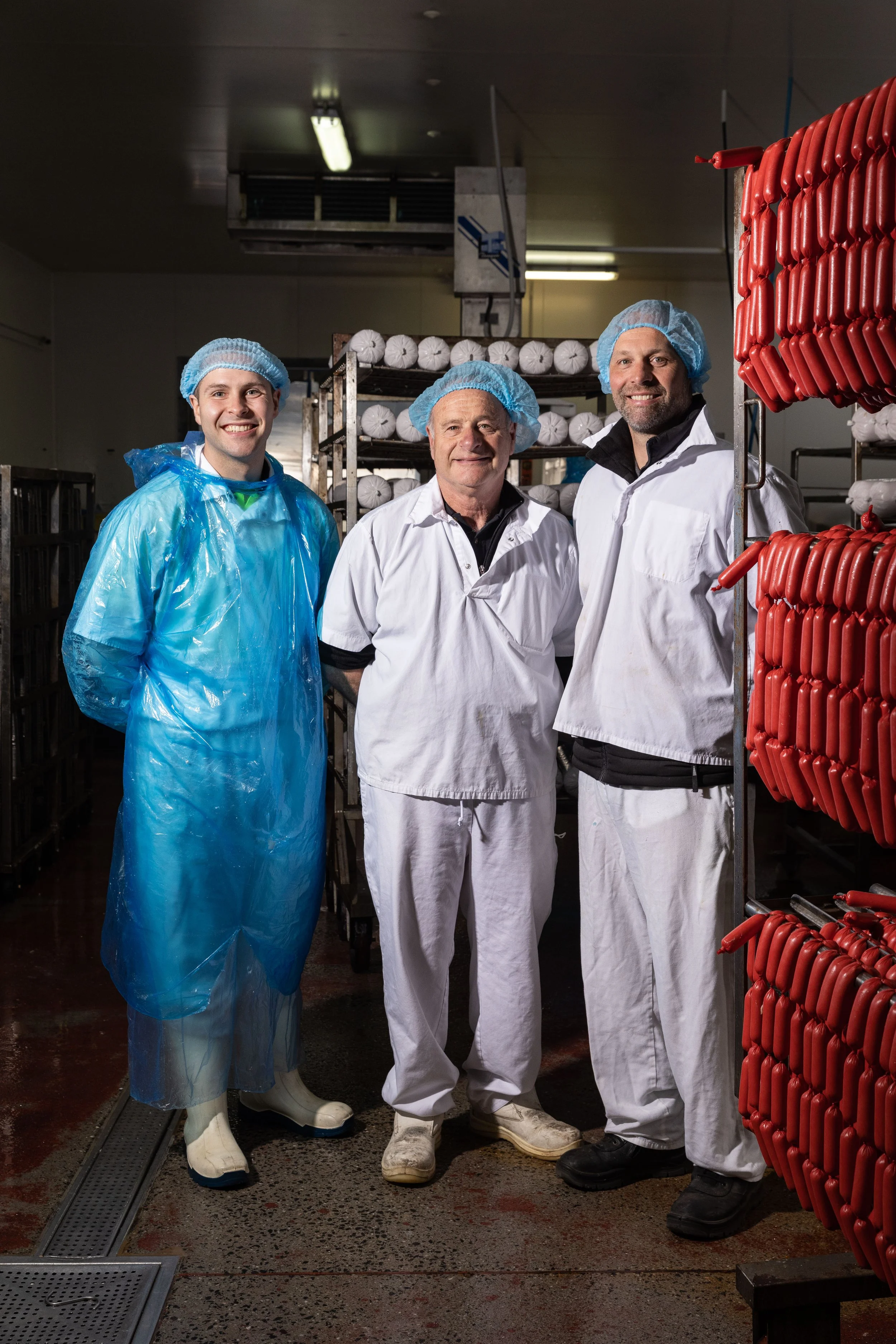 Three men in protective clothing standing in a meat processing facility, surrounded by racks of sausages and meat products.