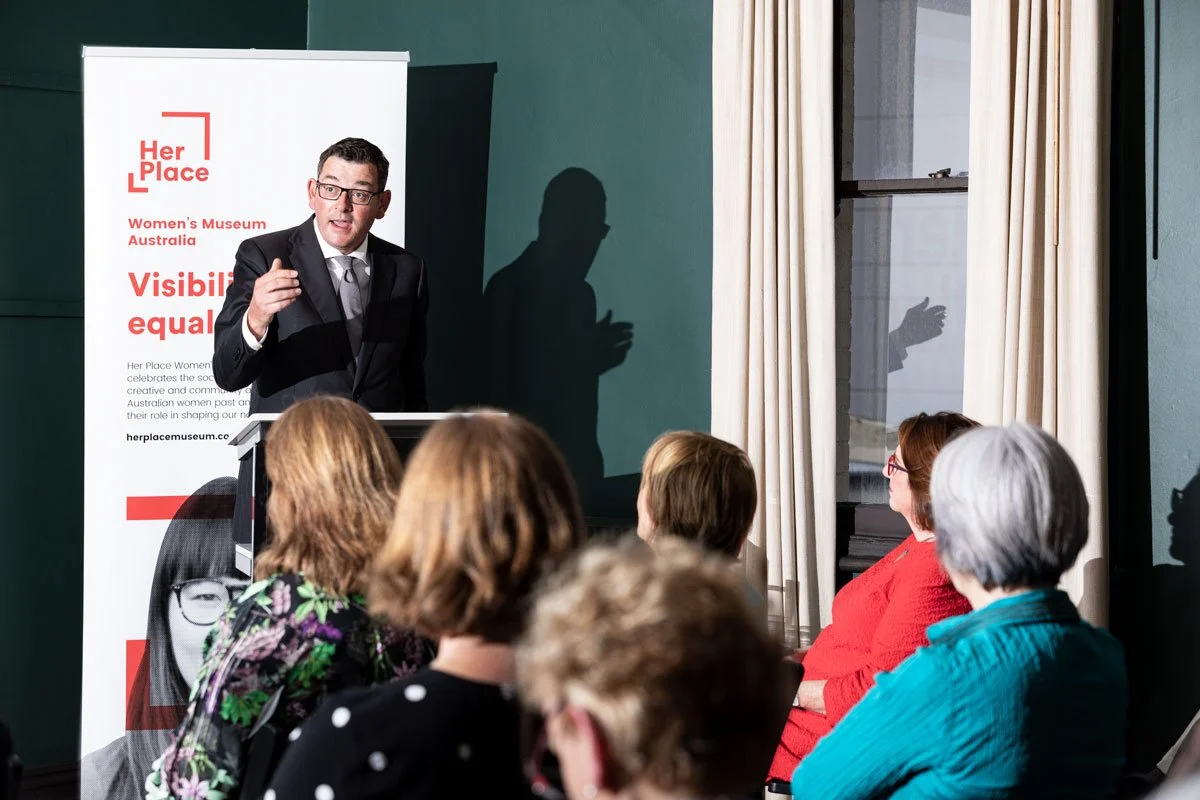 A man in a suit and glasses giving a presentation to a group of women seated in front of him. A banner reads "Her Place, Women's Museum Australia, Visibility equals equality."