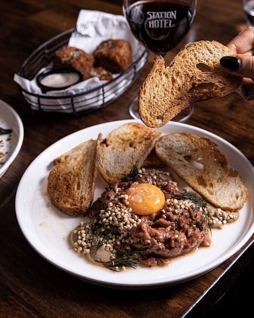 A plate of steak tartare with a raw egg yolk, toasted bread slices, a small bowl of mayonnaise, and a glass of red wine on a wooden table.