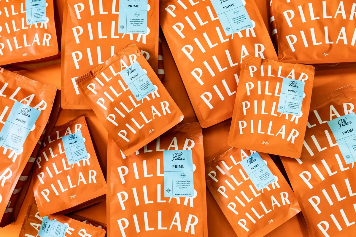 Multiple orange bags of Pillar coffee labeled 'Pillar Prime' with white labels, arranged in a scattered pattern.