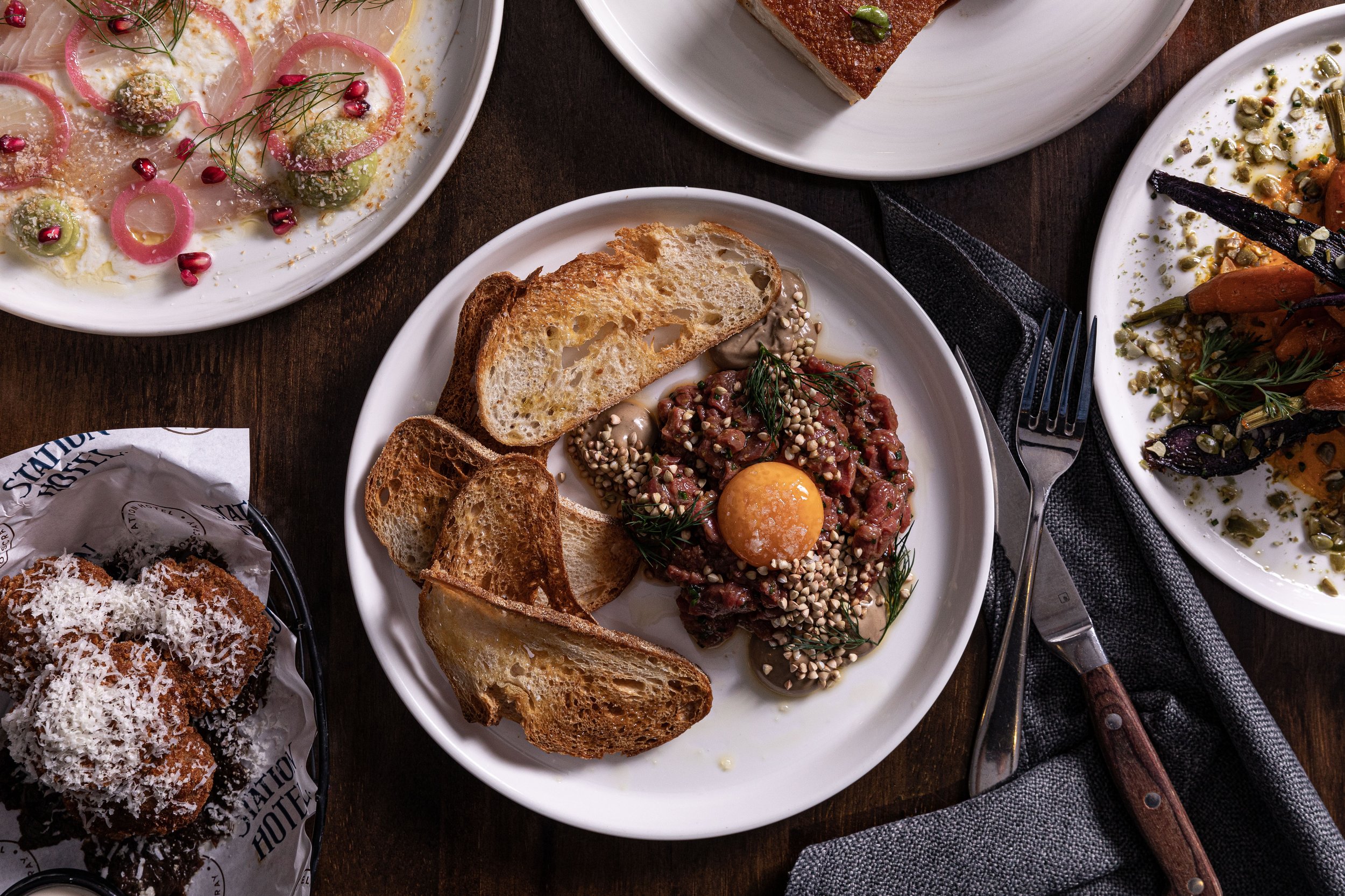 Plate with toasted bread slices and steak tartare topped with an egg yolk, garnished with herbs and sesame seeds, surrounded by various salad dishes and fried food on a wooden table.