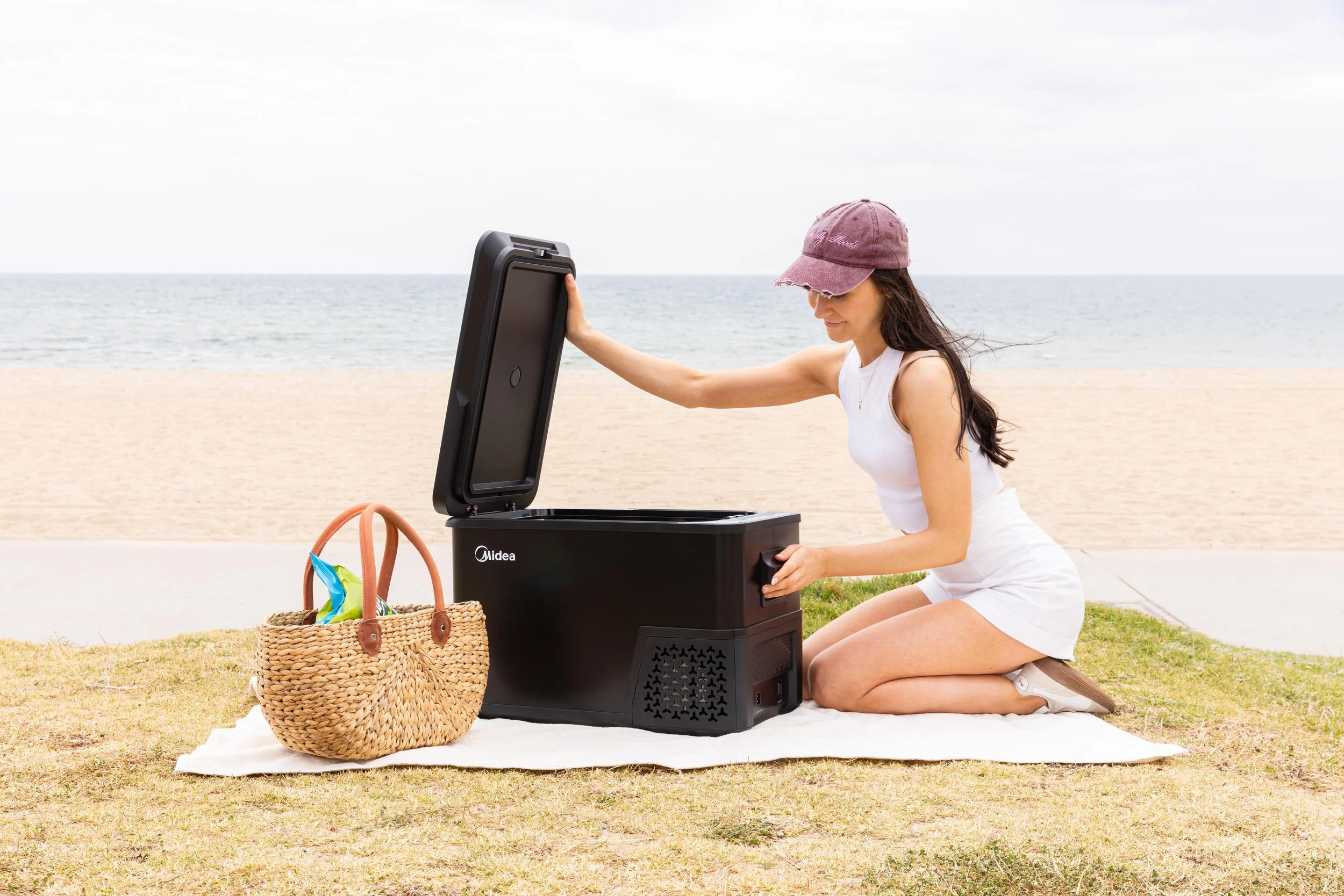 Woman kneeling on a towel on the beach opening a Black Midea portable cooler, with a woven bag nearby holding beach towels and a swim ring.