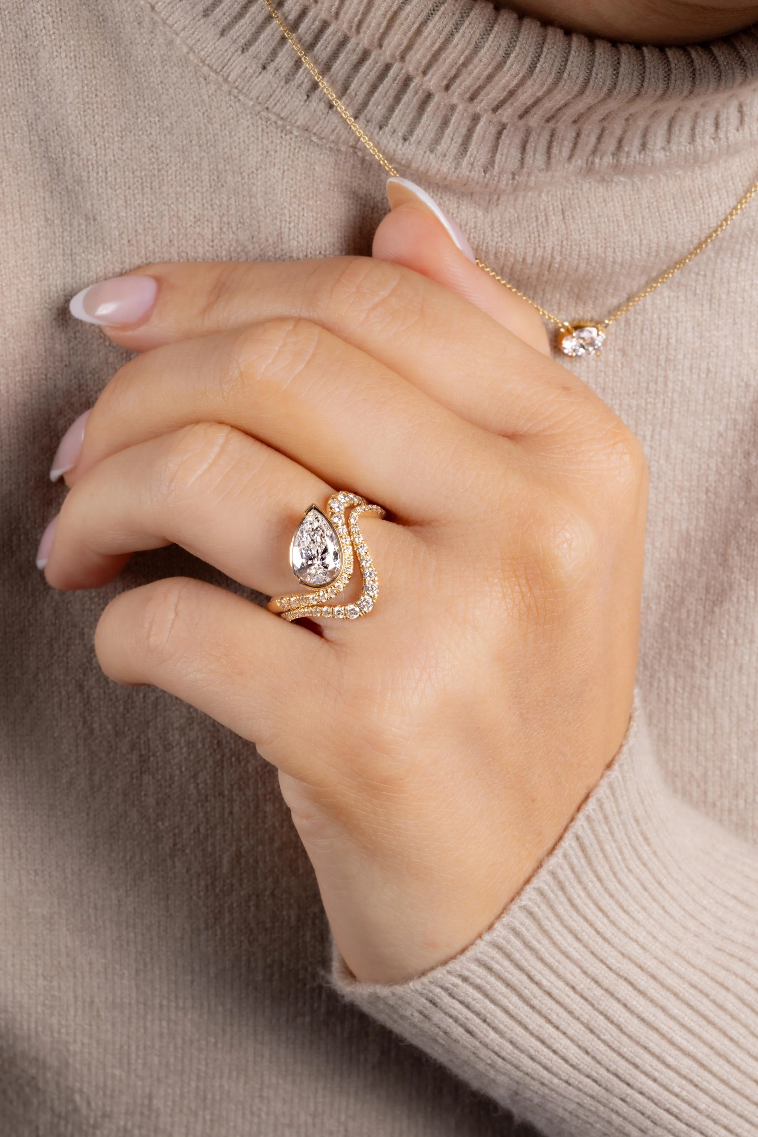 A woman wearing a beige sweater, holding her hand near her chest, displaying a gold ring with a large pear-shaped diamond and smaller diamonds, and a gold necklace with a small diamond pendant.