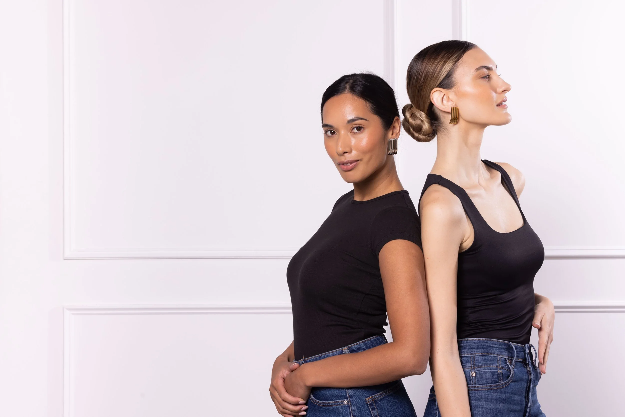 Two women standing back to back, wearing black sleeveless tops and jeans, with their hands on their hips, against a white wall background.