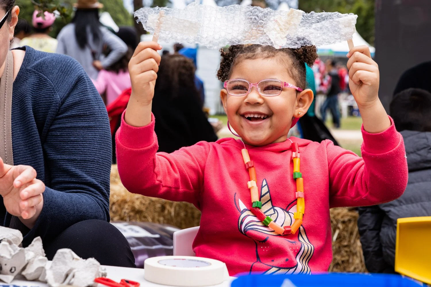 A young girl with curly hair, wearing pink glasses and a red shirt with a unicorn graphic, is smiling and holding a piece of bubble wrap over her head at an outdoor event. She is wearing colorful bead necklaces and is seated beside an adult.