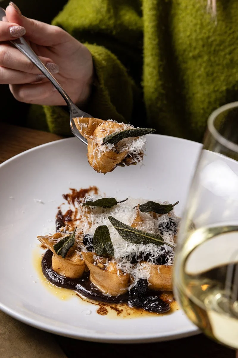 Close-up of a fork holding a piece of pasta with black truffle and sage leaves, over a white plate of pasta garnished with grated cheese, sage leaves, and dark sauce, with a glass of white wine partially visible in the foreground.
