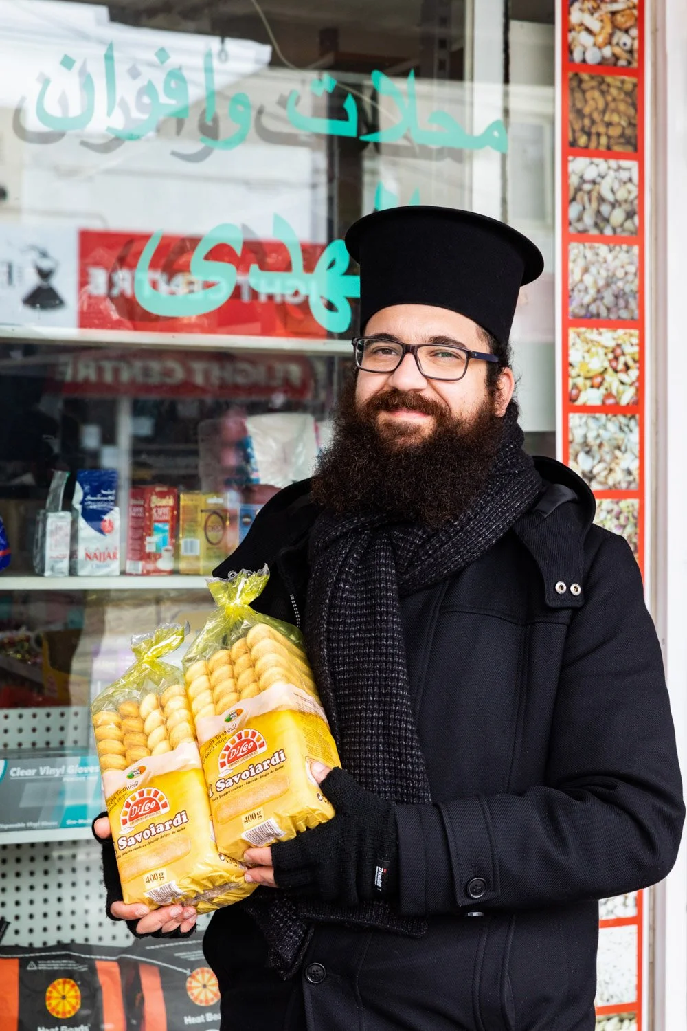 A man with a beard, glasses, wearing a black hat, coat, and scarf, holding two bags of pasta outside a shop with a reflective window.