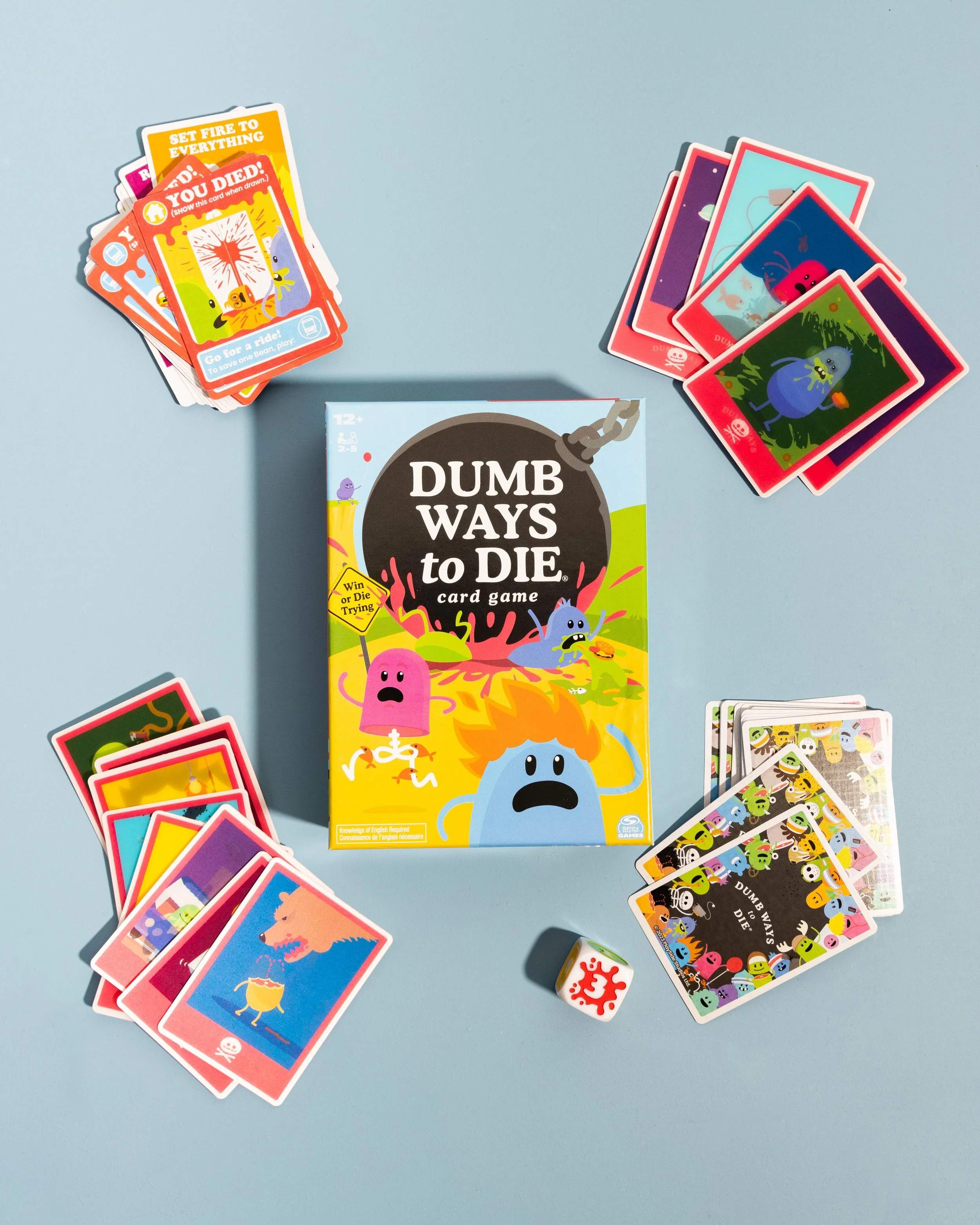 Dumb Ways to Die card game box on a light blue surface surrounded by colorful playing cards and a white die with a red splash pattern.