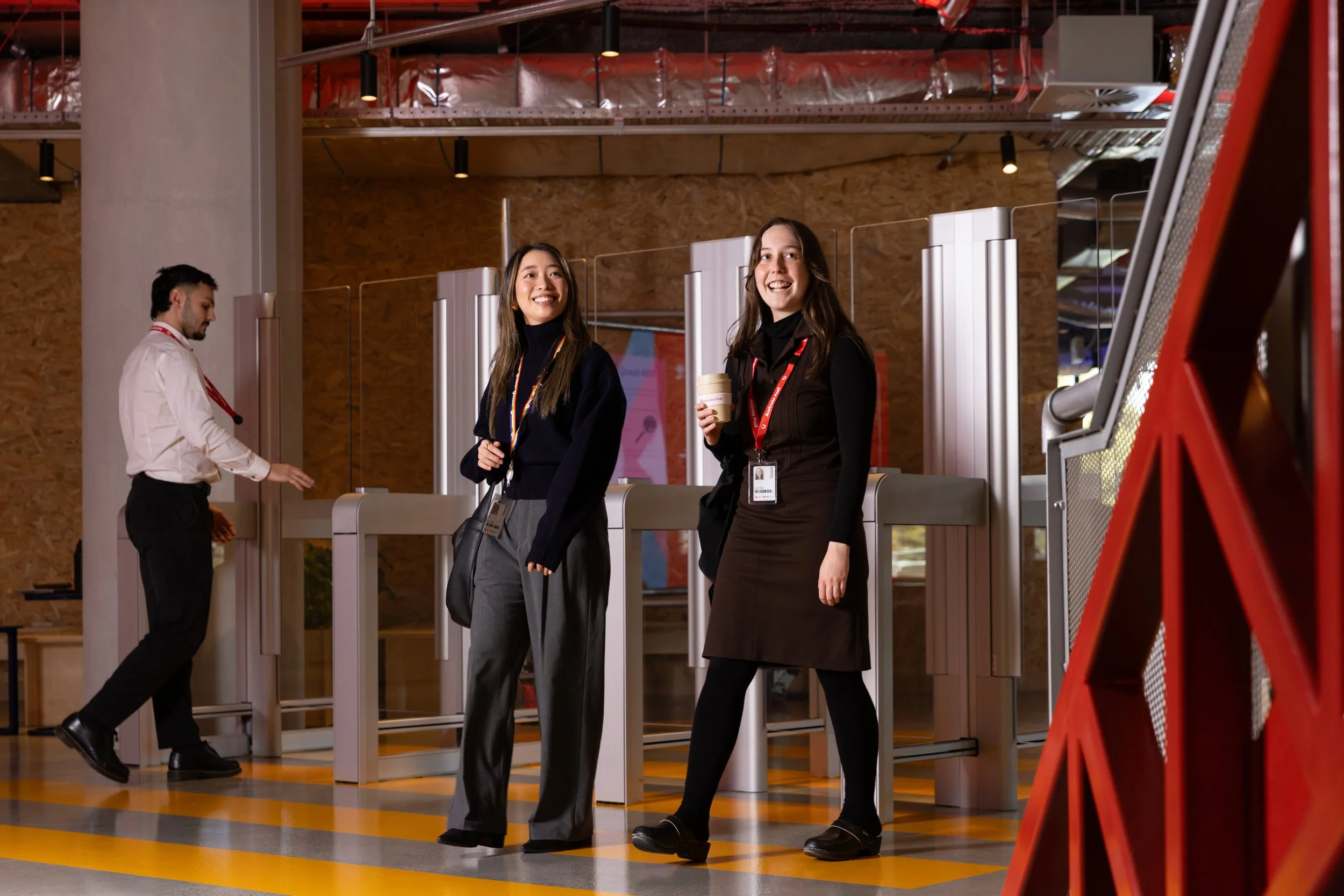 Two women smiling and standing near security turnstiles in an office building lobby, one holding a coffee cup, with a man checking his phone in the background.