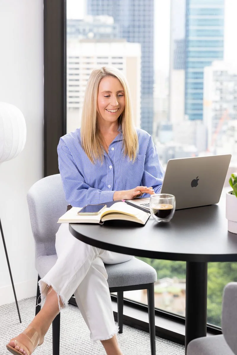 A woman with blonde hair sitting at a round table in an office with large windows, working on a laptop, with a glass of coffee, an open notebook, and a smartphone on the table.