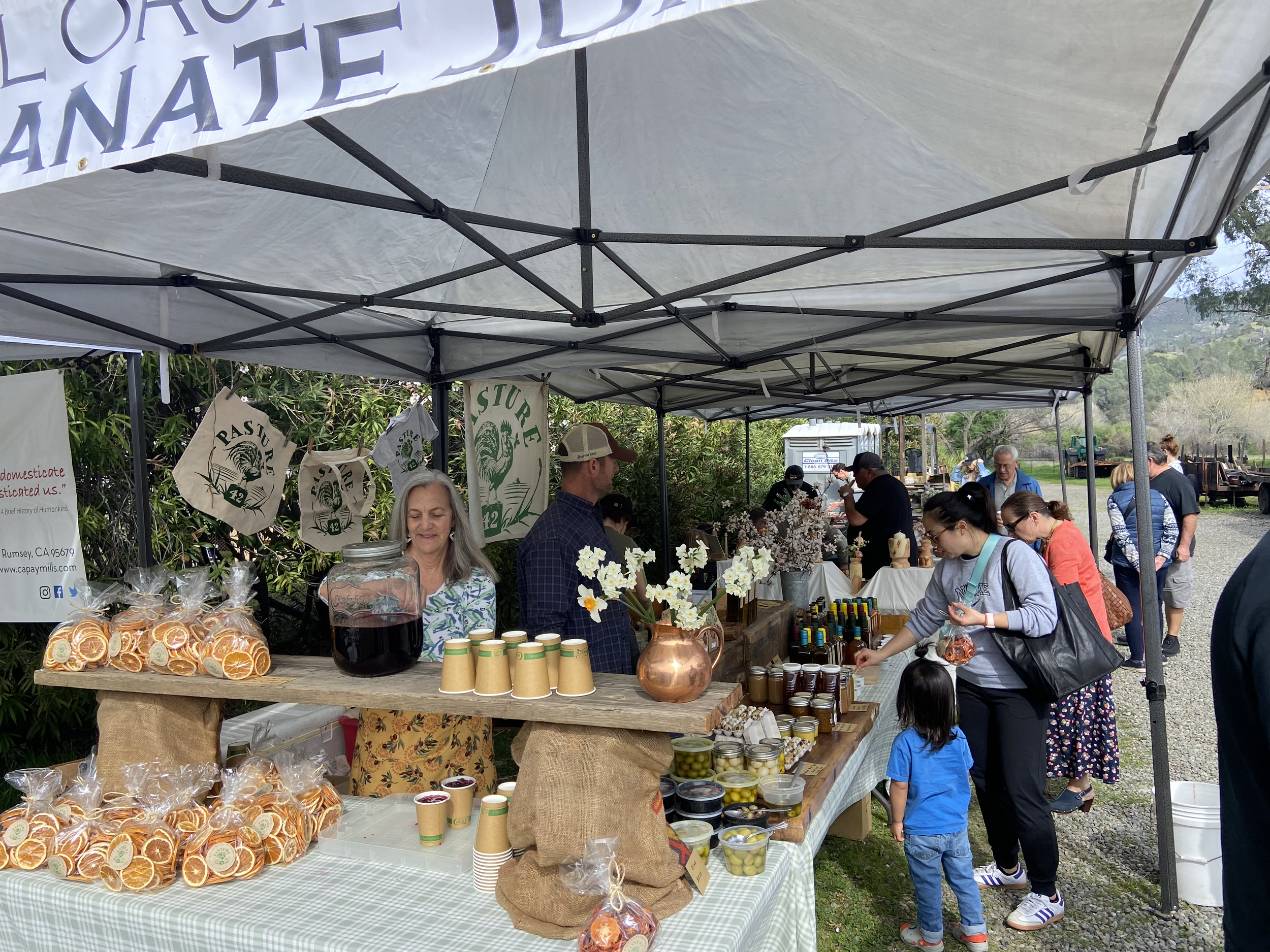 People shopping at an outdoor farmers market stall with homemade goods, jams, and dried oranges, under a white canopy with signs that read 'FLOURISH' and 'NATURAL'.