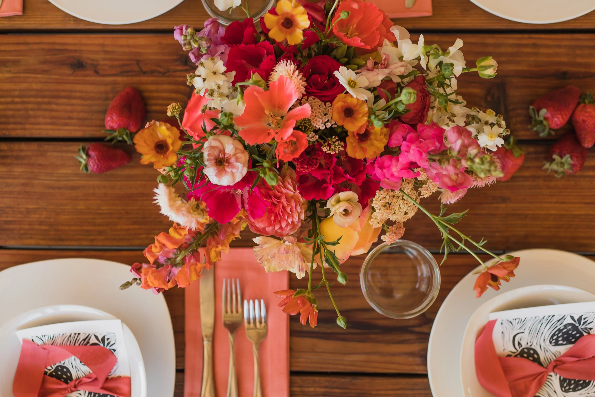 Top view of a table setting with a large colorful flower arrangement, strawberries, white plates with black and white napkins tied with pink ribbons, and gold cutlery on a pink cloth napkin, on a wooden table.