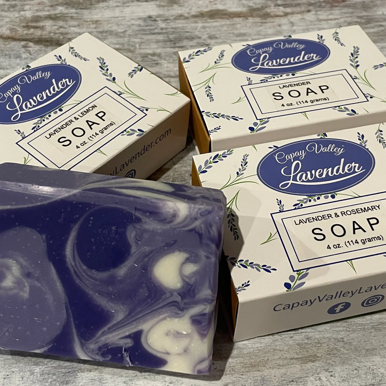 Three boxes of Capay Valley Lavender soap and a purple and white marbled soap bar on a wooden surface.