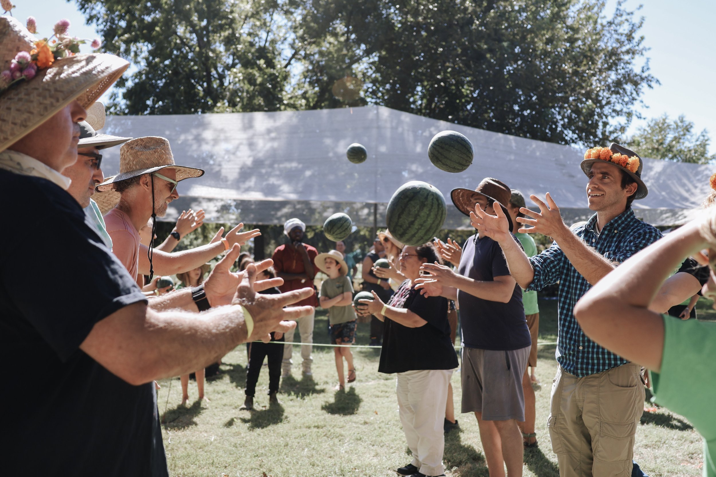 People wearing hats and casual clothes participating in a watermelon juggling game outdoors during the day.