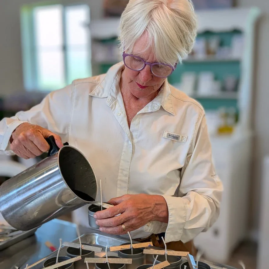 An elderly woman with glasses and white hair pours liquid into small black containers in a kitchen or laboratory setting.