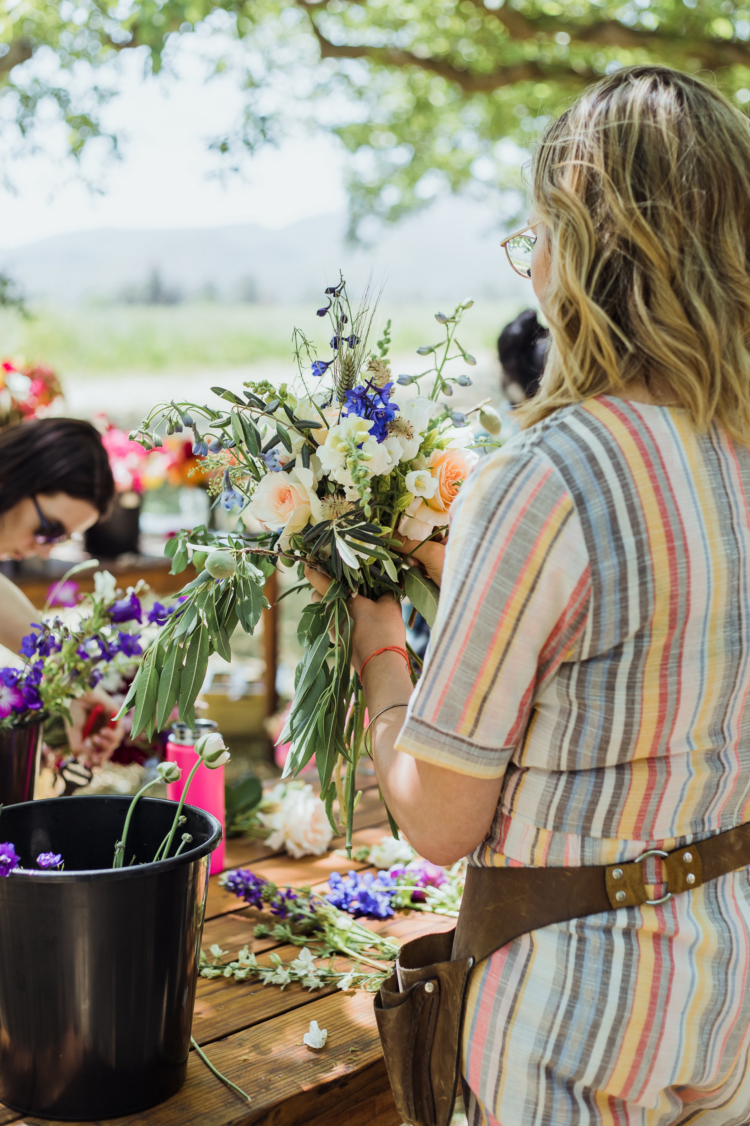 Woman arranging a bouquet of flowers outdoors at a workshop, with other people working on floral arrangements at a wooden table under a tree.
