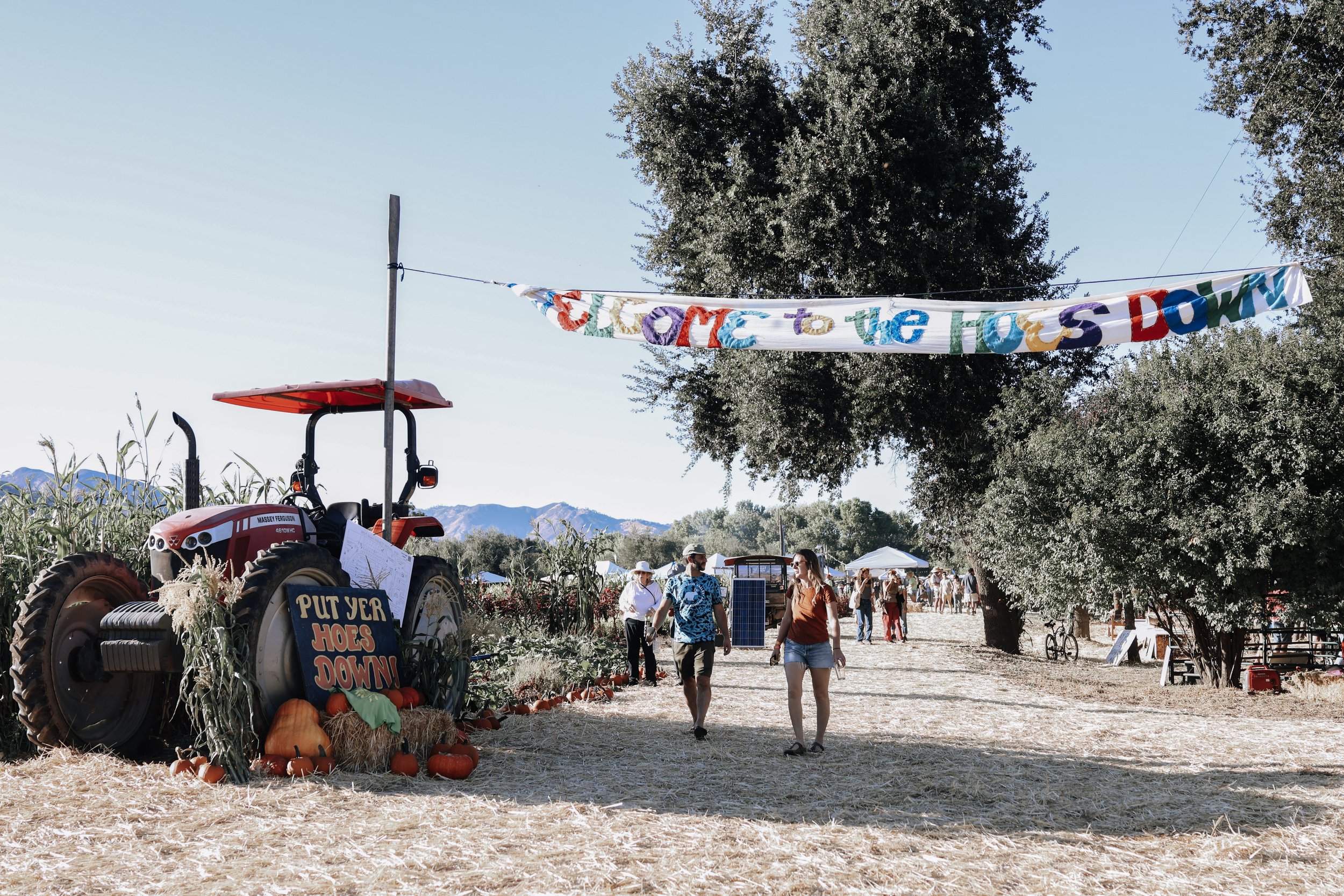 People walking through an outdoor festival with a large sign that says 'Clore to the Hoes DOWN' hanging from trees. There is a red tractor decorated with pumpkins and fall decor, and a colorful banner that reads 'PUT YOUR HOES DOWN.' In the background, there are tents, trees, and mountains.