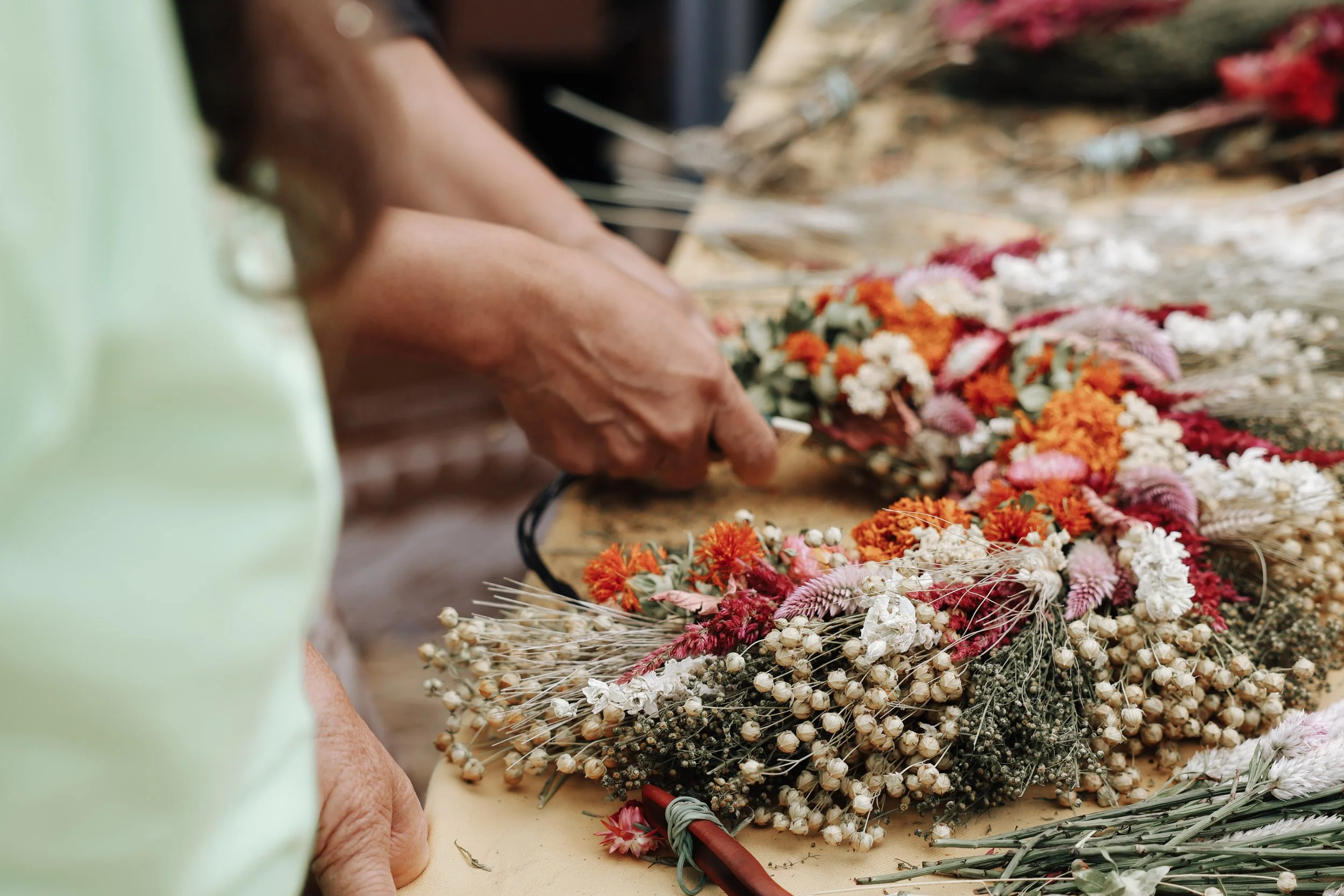 Close-up of a person's hands assembling a floral arrangement with various colorful dried flowers and plants on a wooden surface.