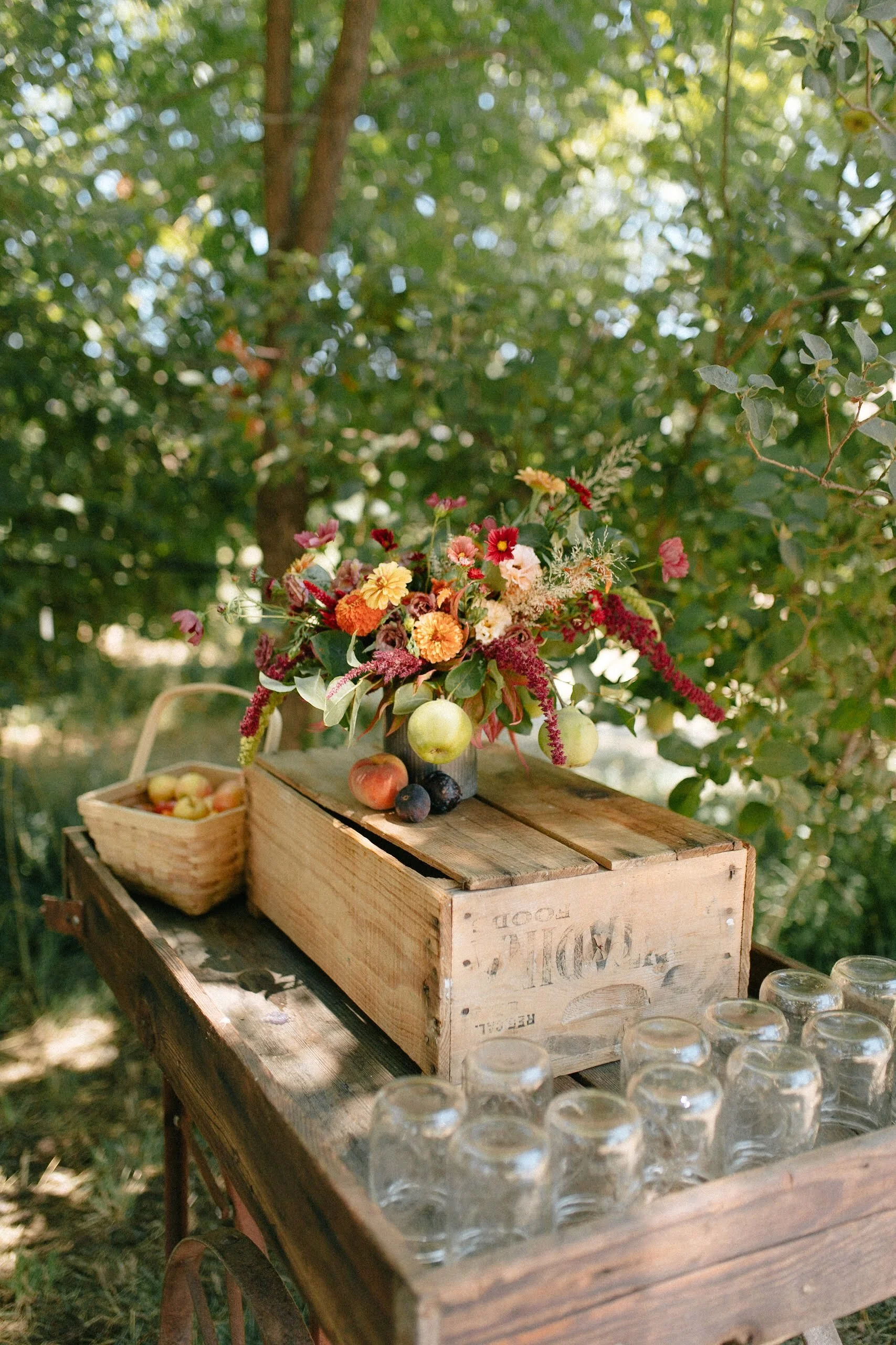 A rustic outdoor table with a wooden crate, filled with a colorful flower arrangement, apples, and grapes, with a basket of apples and upside-down glasses nearby, set in a lush, green, sunlit garden.