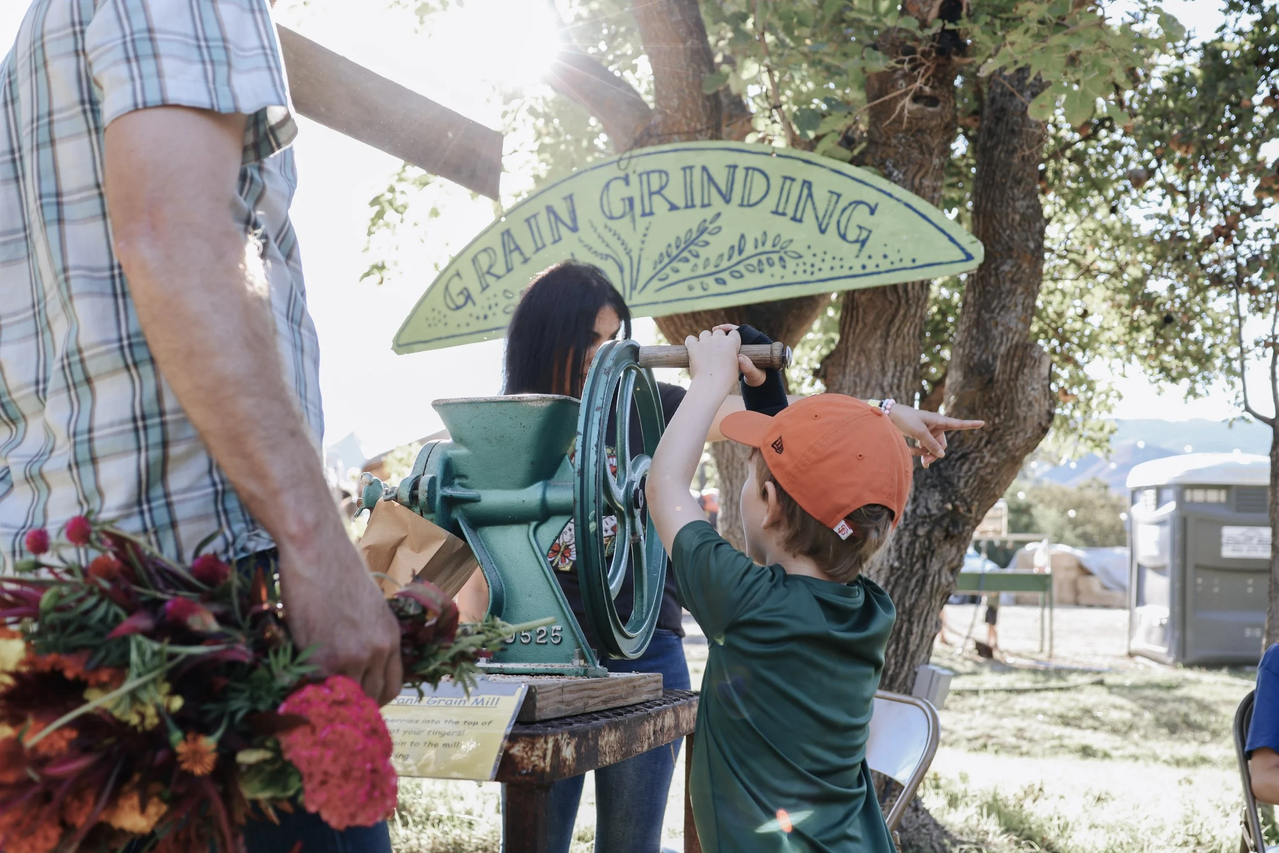 Child operating a manual grain grinder at a farm booth, with a woman and a person holding flowers nearby, under a tree with a sign reading 'GRAIN GRINDING' in the background.