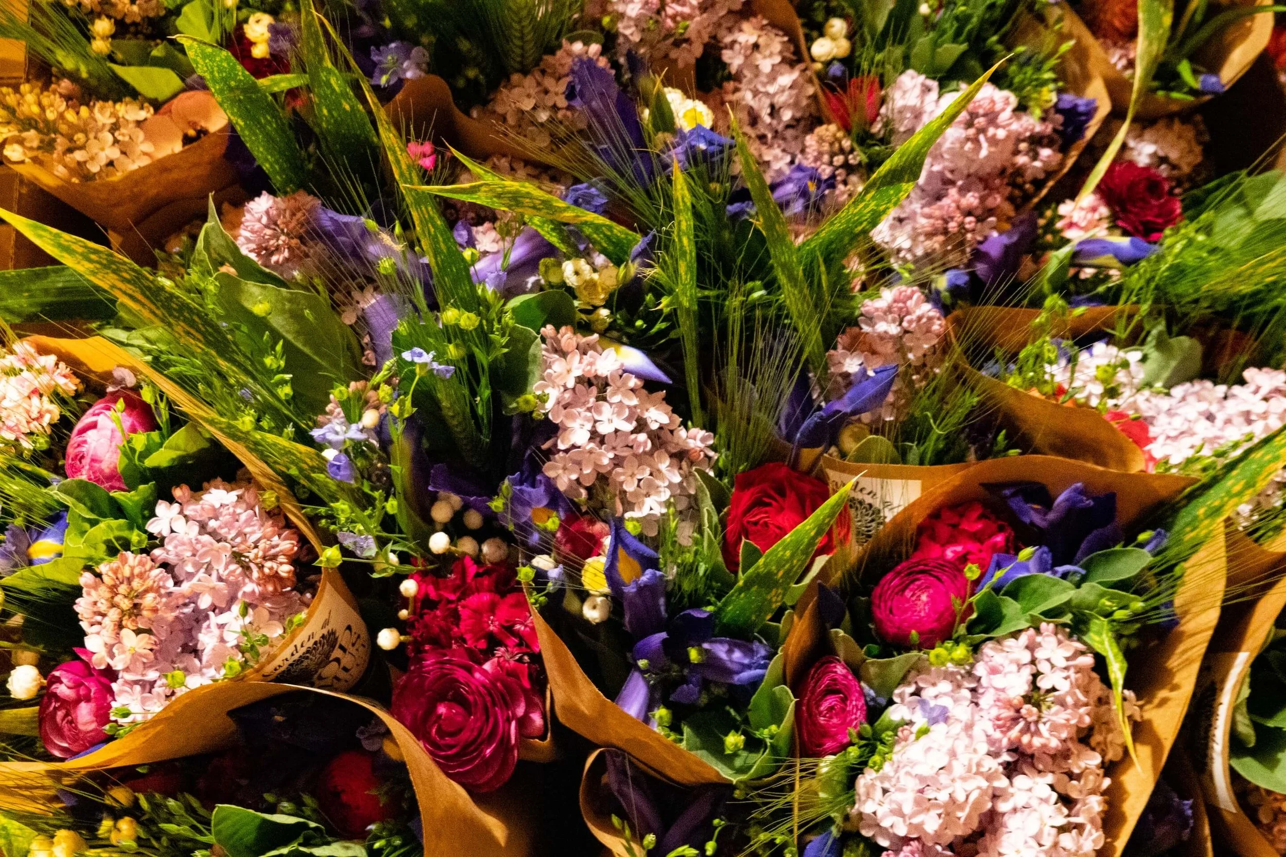 Bouquets of assorted colorful flowers including pink, purple, red, and white blooms, wrapped in brown paper with green foliage and leaves.