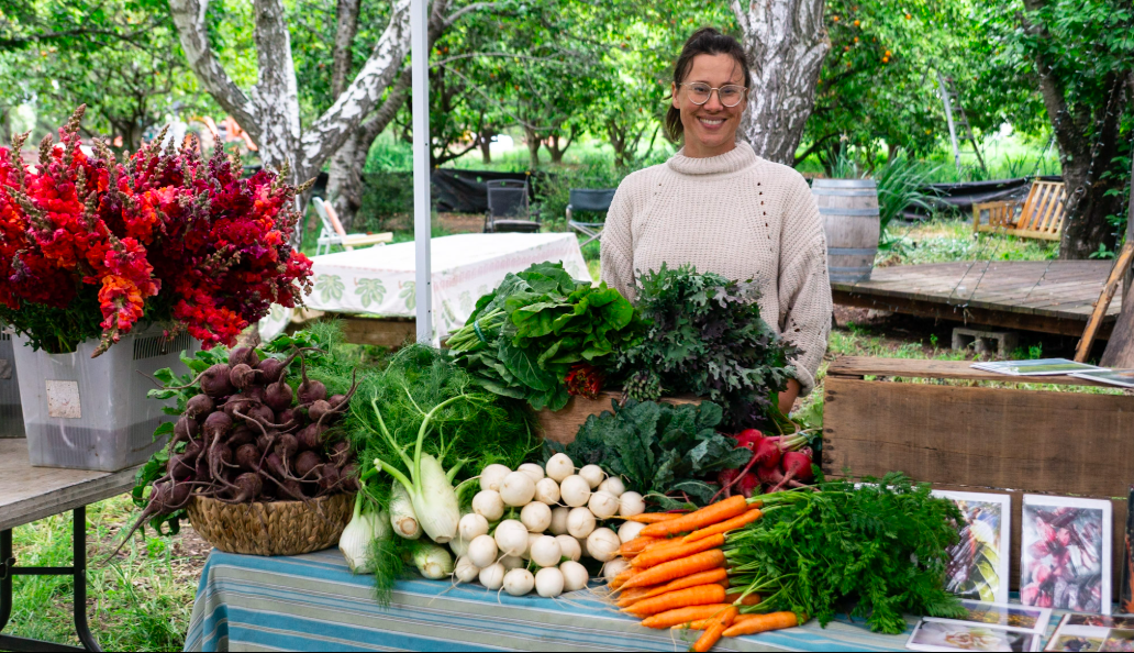 A woman standing behind a table at a farmer's market stall displaying fresh vegetables and flowers, with trees and a wooden deck in the background.