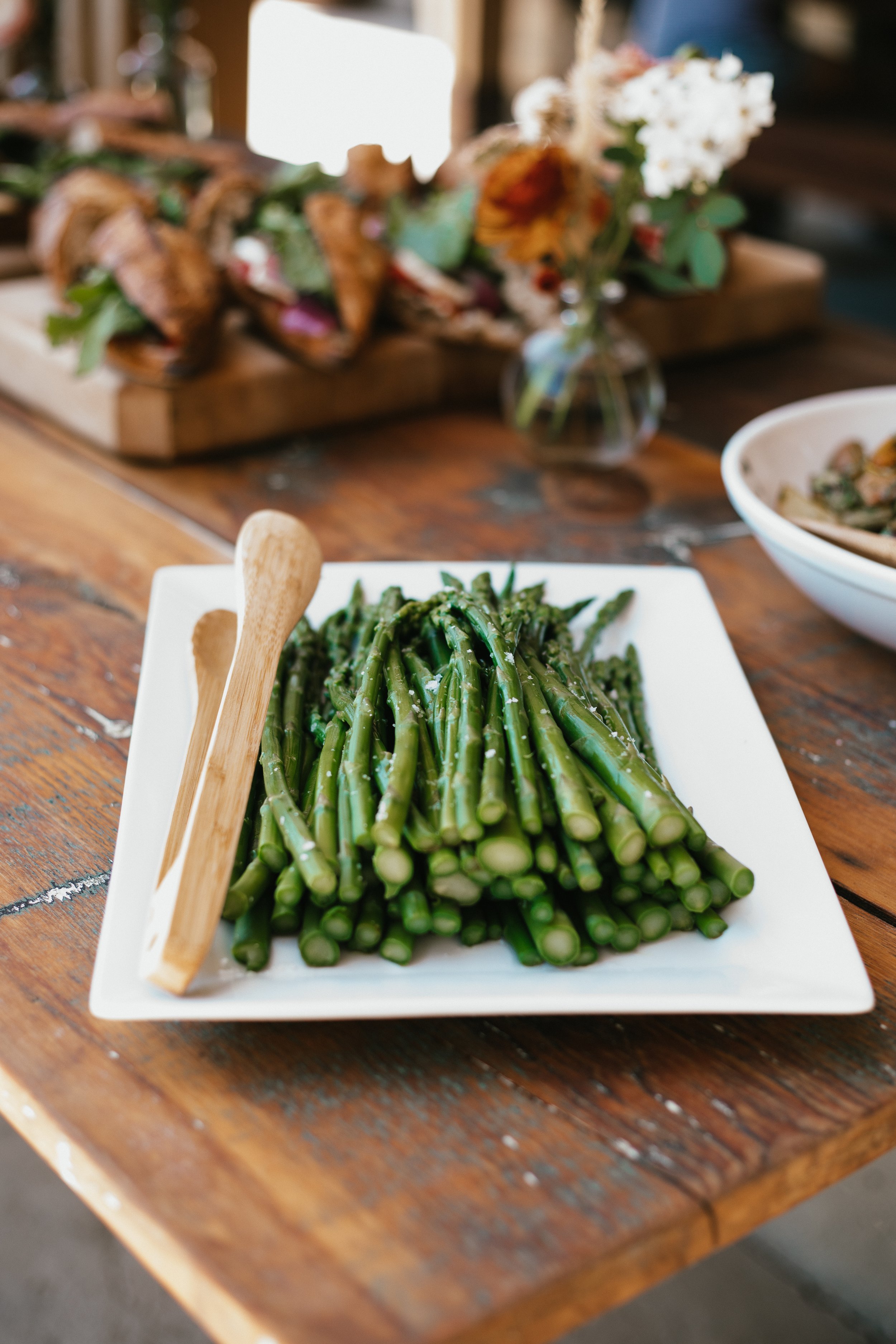 Plate of cooked green asparagus spears on a white square dish with wooden tongs, on a rustic wooden table.
