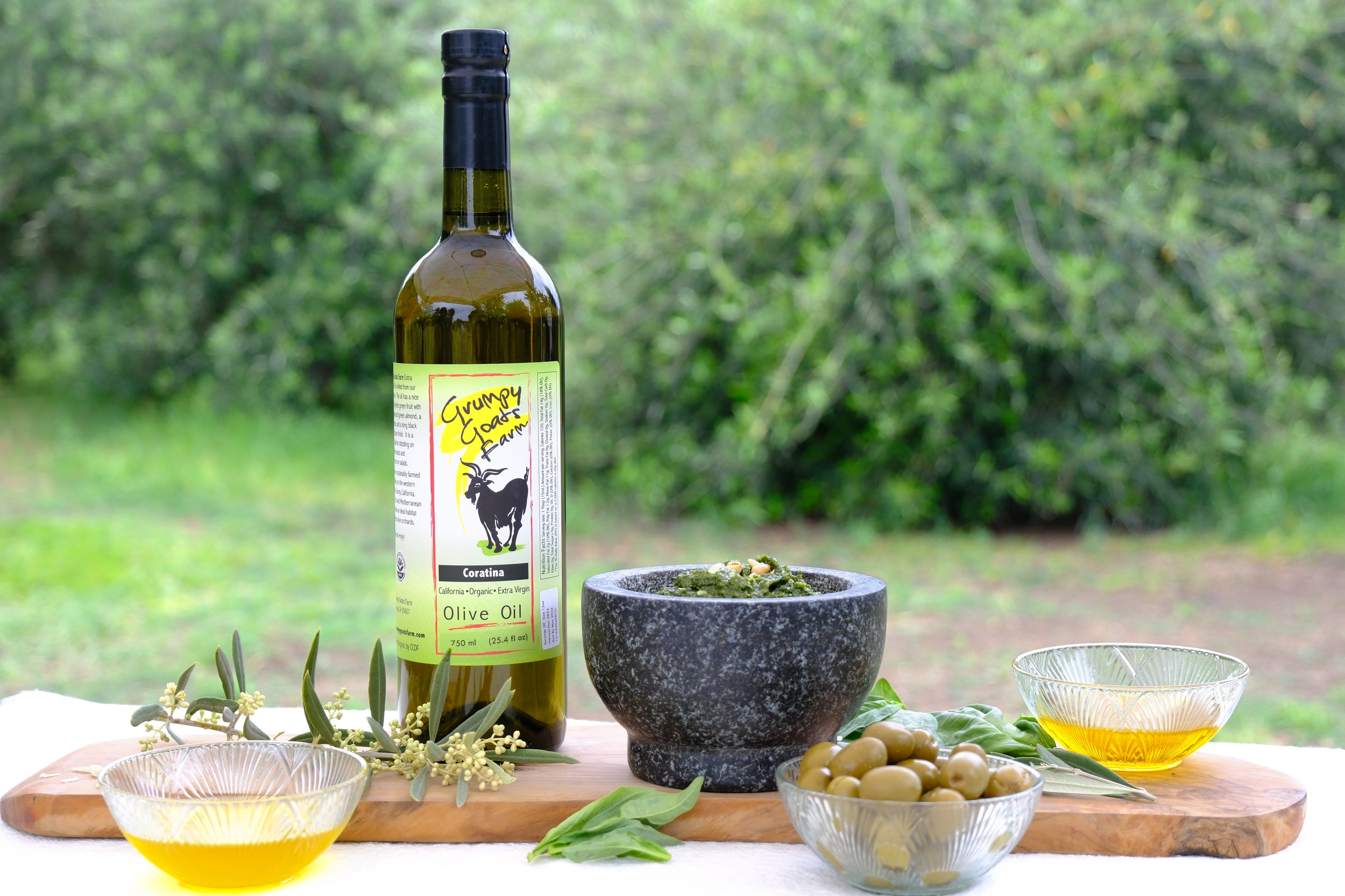 A bottle of Grumpy Goat Farms extra virgin olive oil, a bowl of green olives, a small bowl of olive oil, and a mortar and pestle containing a green pesto, all arranged on a wooden board outdoors with a blurred green background.