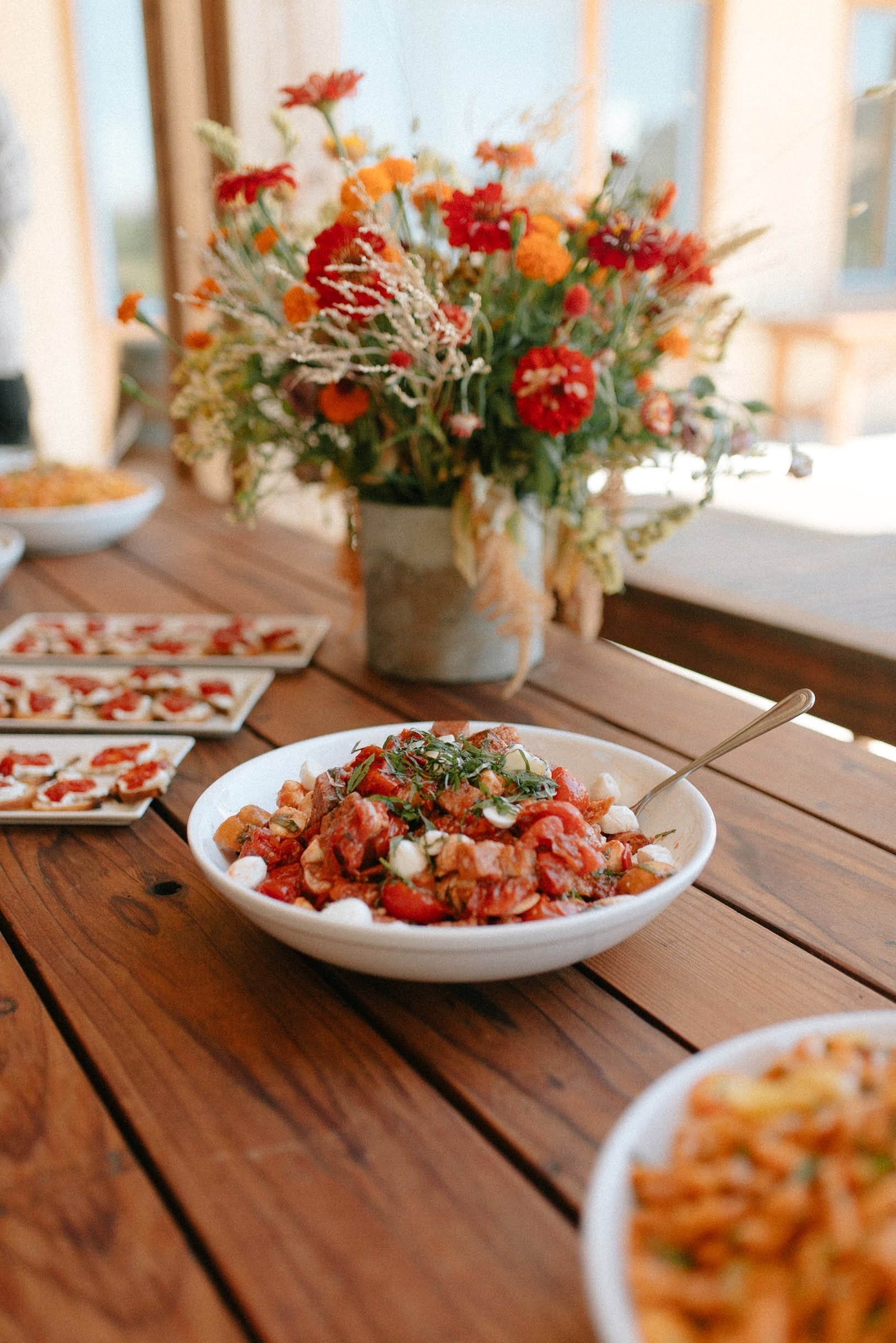 A wooden table with a bowl of tomato and vegetable salad, a spoon, and small plates of appetizers, with a vase of colorful flowers in the background.