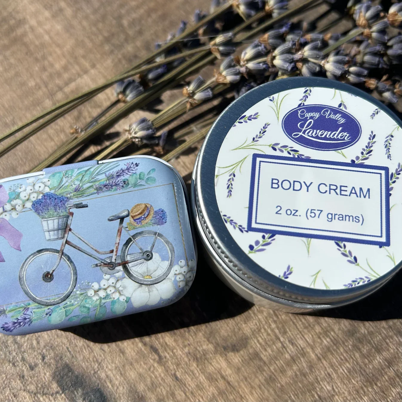 A round container of Capay Valley Lavender body cream and a decorative tin featuring a bicycle with a basket of lavender and white flowers on a wooden surface with dried lavender flowers in the background.