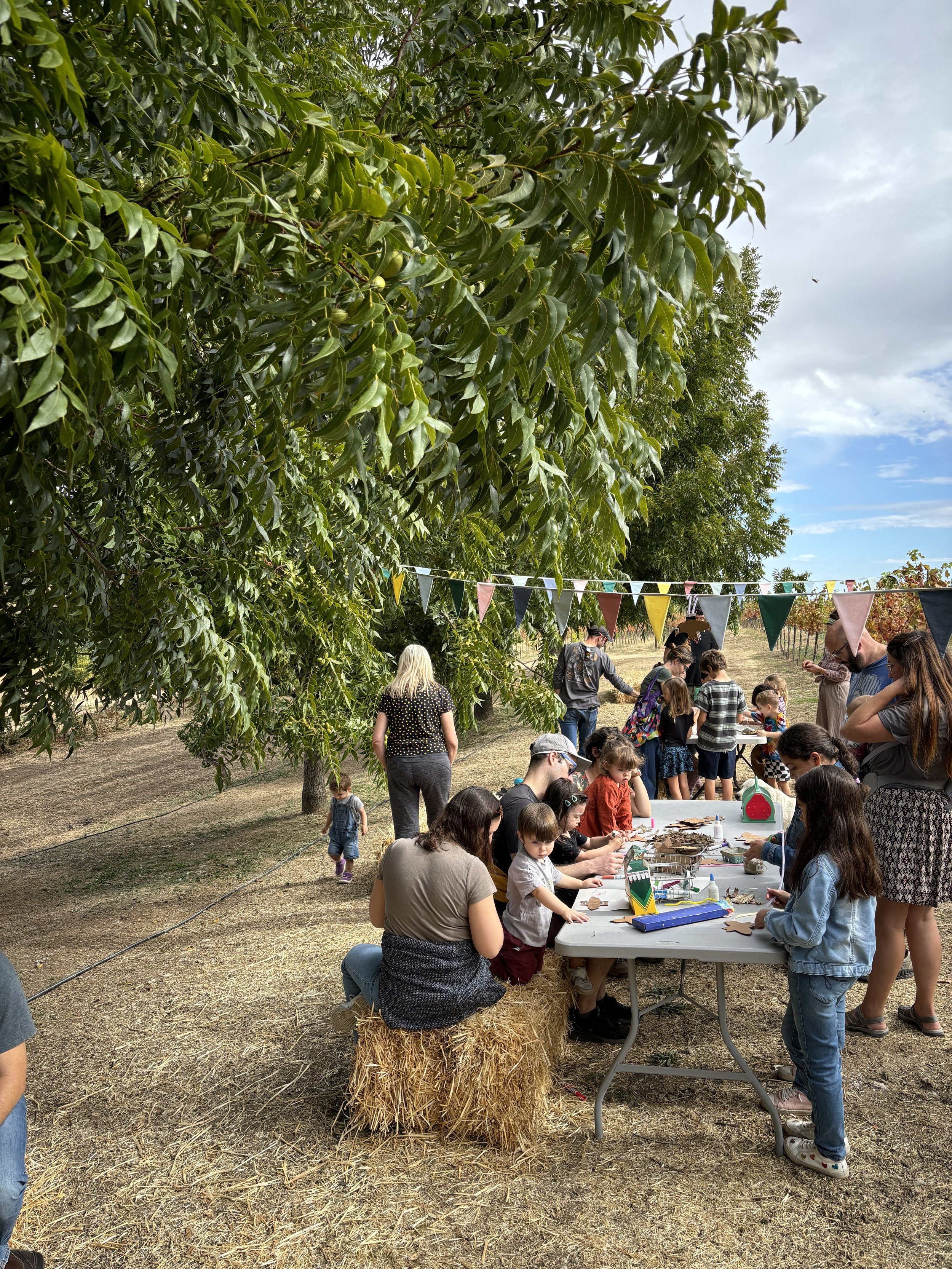People gathered outdoors at a craft activity under a large tree with colorful bunting, kids and adults participating, some sitting on hay bales, in a rural setting with cloudy sky.