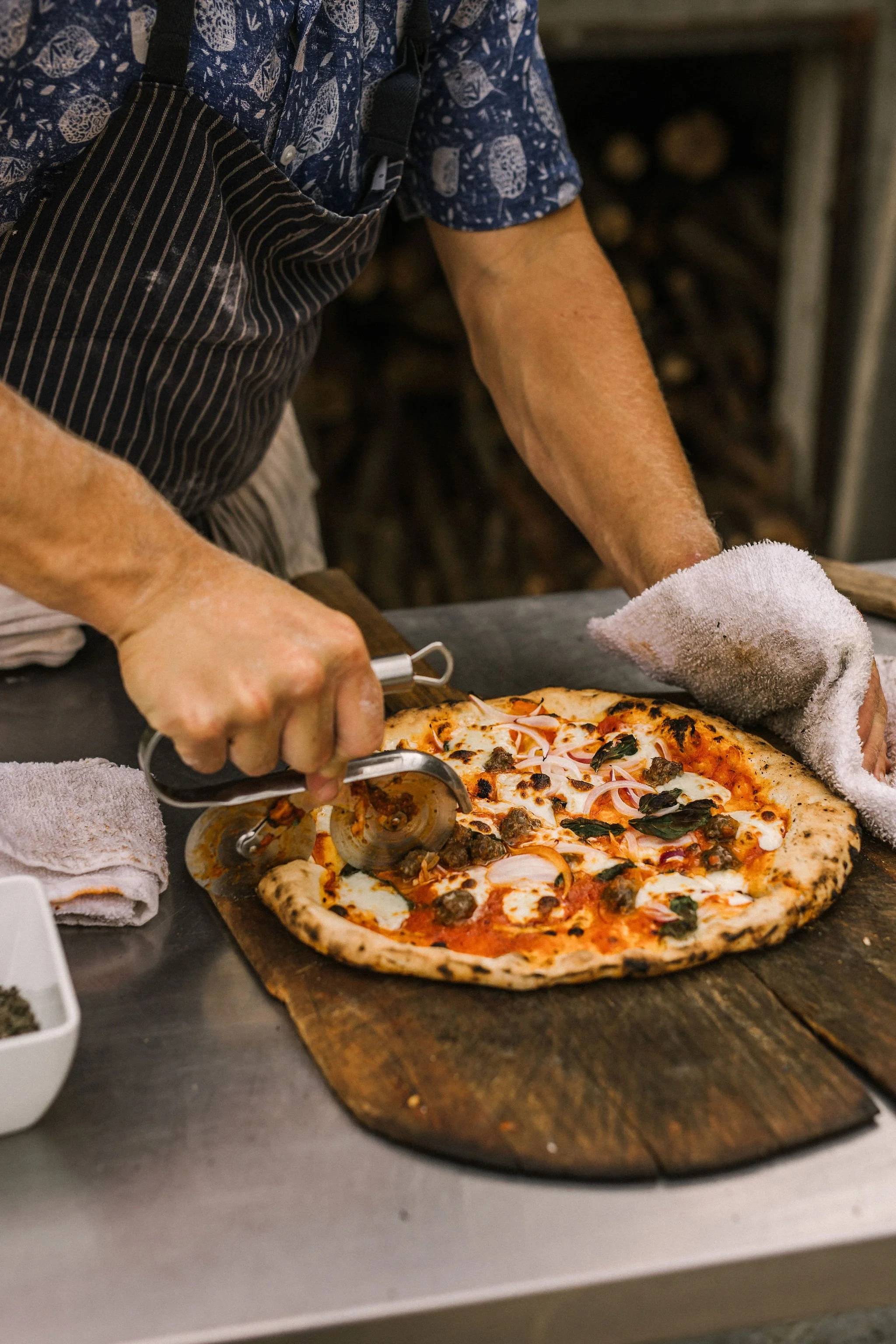 Person slicing a freshly baked pizza topped with cheese, pepperoni, and basil on a wooden paddle, wearing a striped apron and handling a towel.