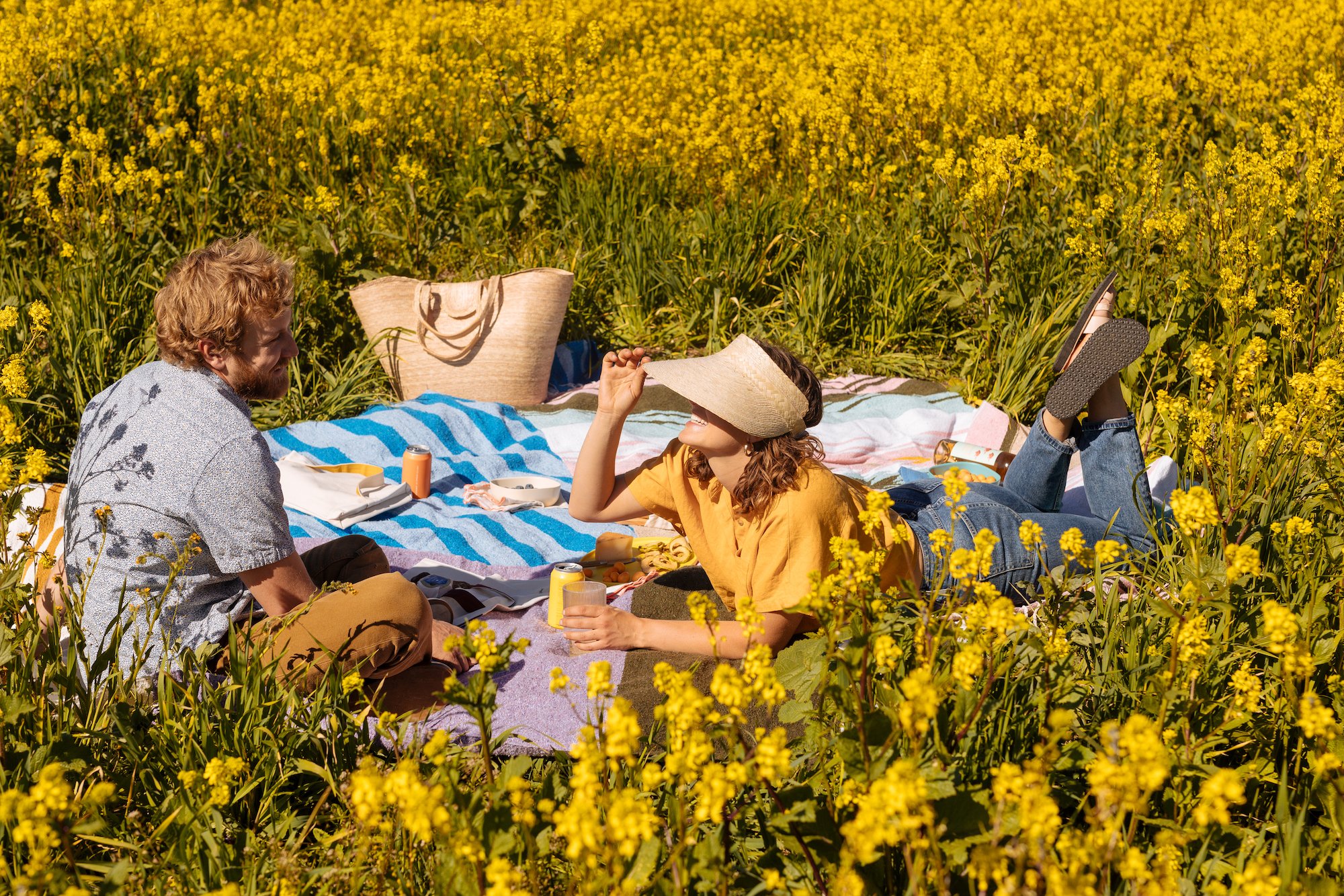 Two people having a picnic in a yellow flower field, sitting on blankets, with drinks and snacks.