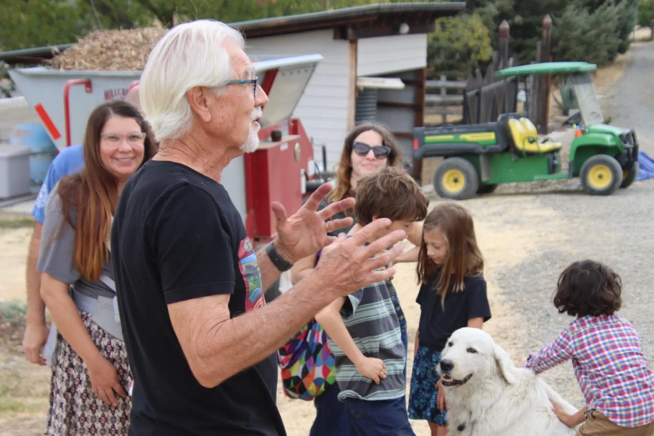 A group of children and adults gathered outdoors around an elderly man with white hair and glasses, speaking with animated hand gestures. A large white dog sits nearby, and a green golf cart is in the background.