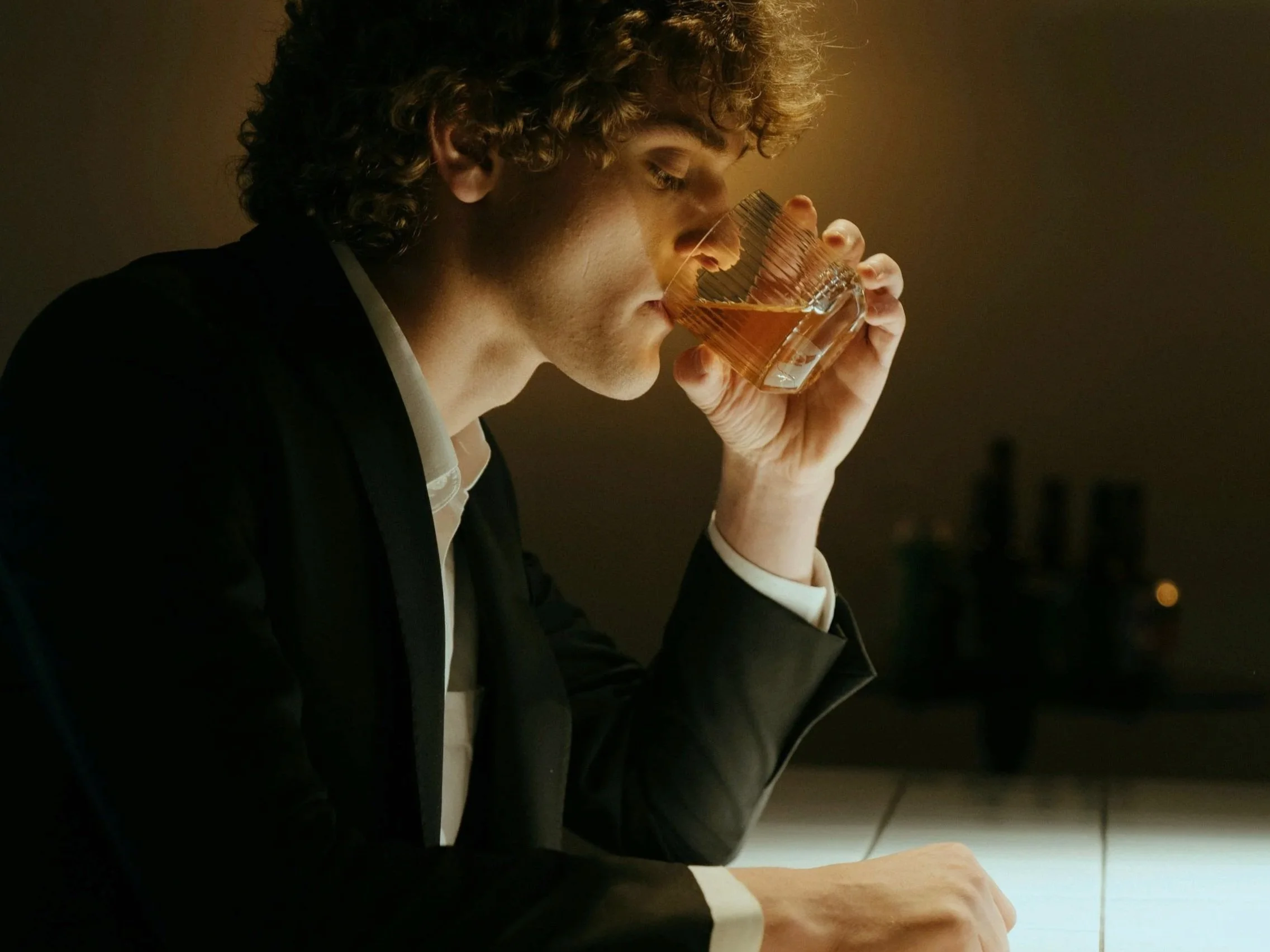 A young man with curly hair in a black suit and white shirt drinking from a glass of whiskey at a dimly lit bar or restaurant.