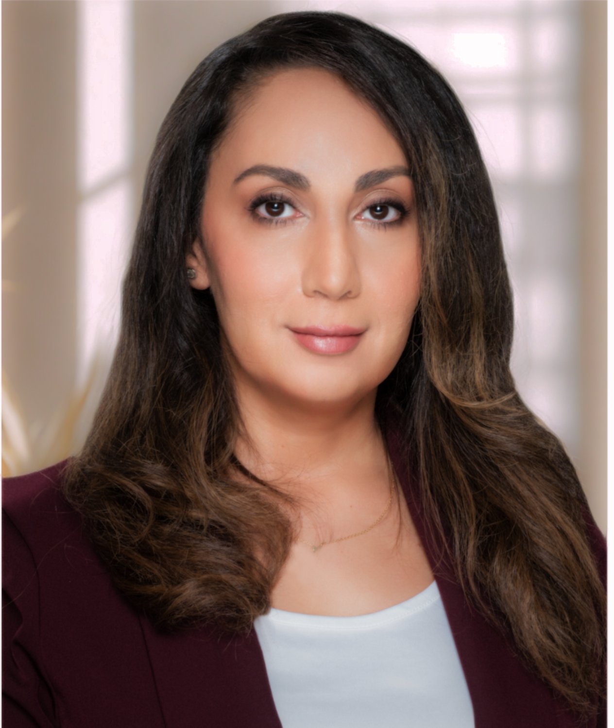 A woman with long dark hair, wearing a maroon blazer and a white top, posing indoors with a blurred background.
