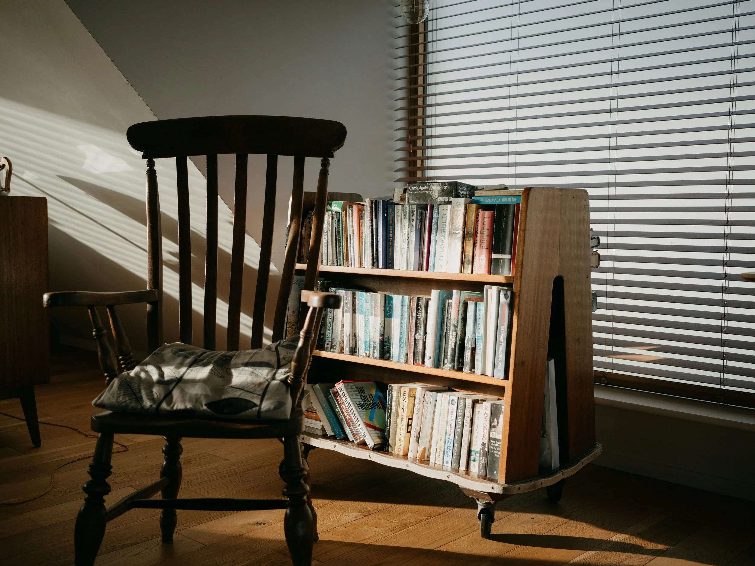 A wooden rocking chair with a cushion placed on it, beside a wooden bookcase filled with books, near a window with closed blinds, sunlight casting shadows inside the room.