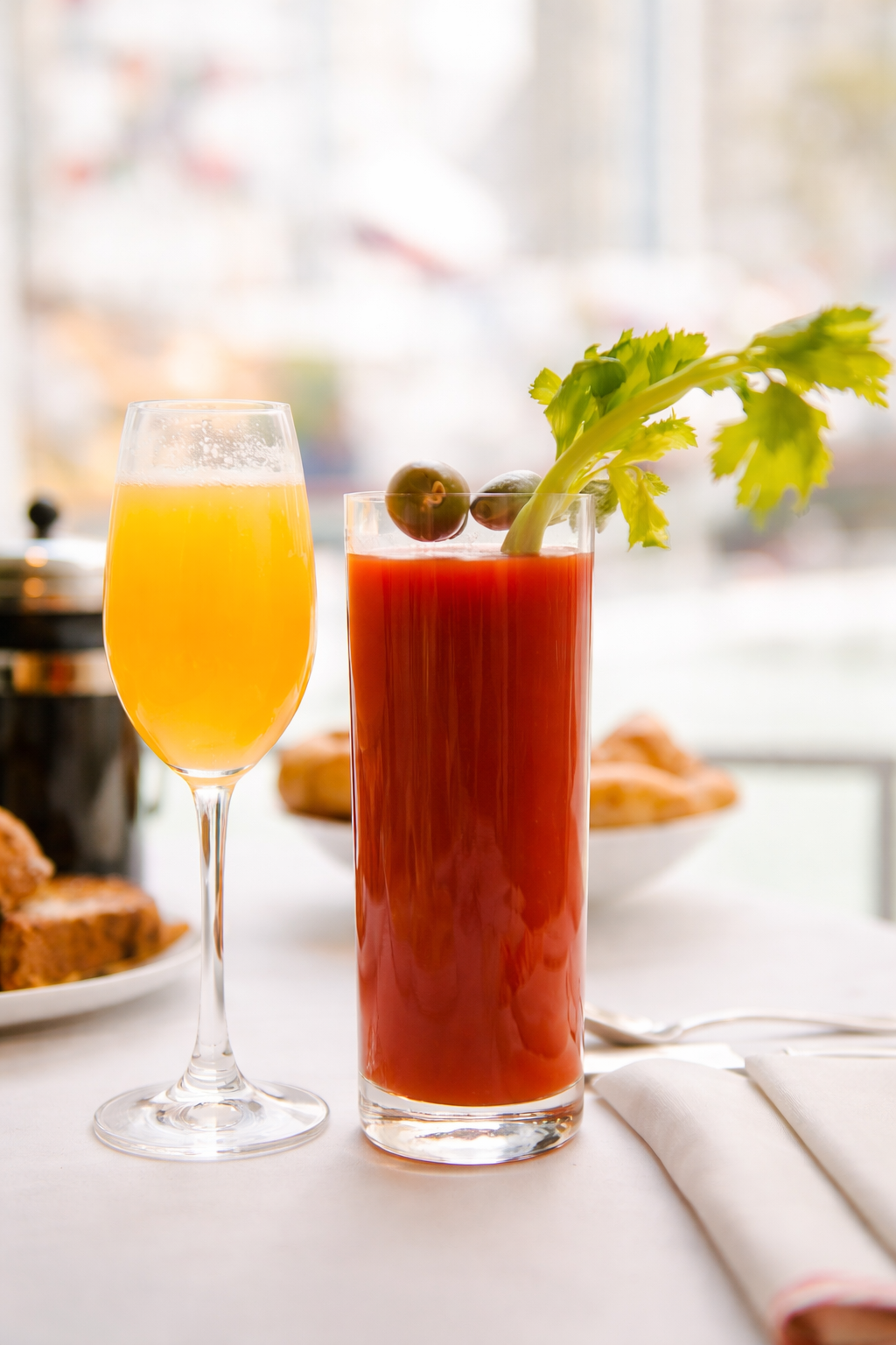 Mimosa in a champagne flute and a bloody mary with olives and celery stalk on a white table with blurred food in the background.