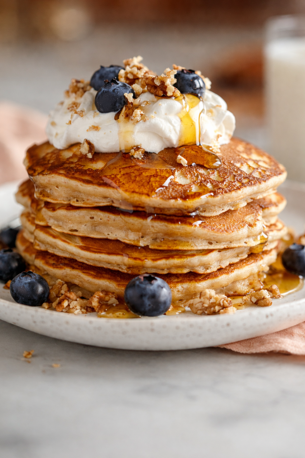 Stack of five pancakes topped with whipped cream, blueberries, chopped nuts, syrup, and a drizzle of honey, on a white plate with blueberries and nuts around it.