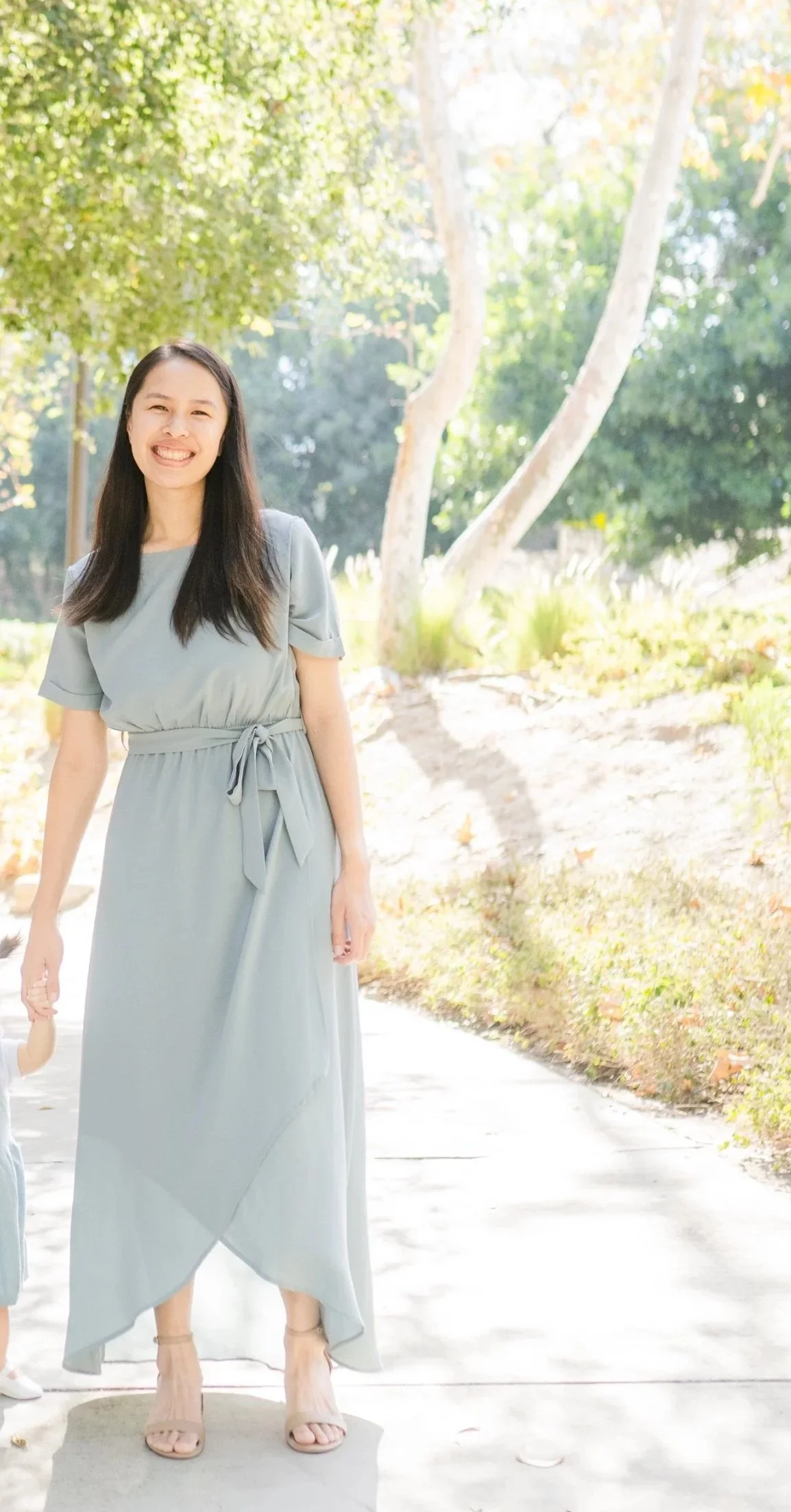 A smiling woman in a light gray dress with a tied waist stands outdoors on a bright sunny day, with trees and greenery in the background.