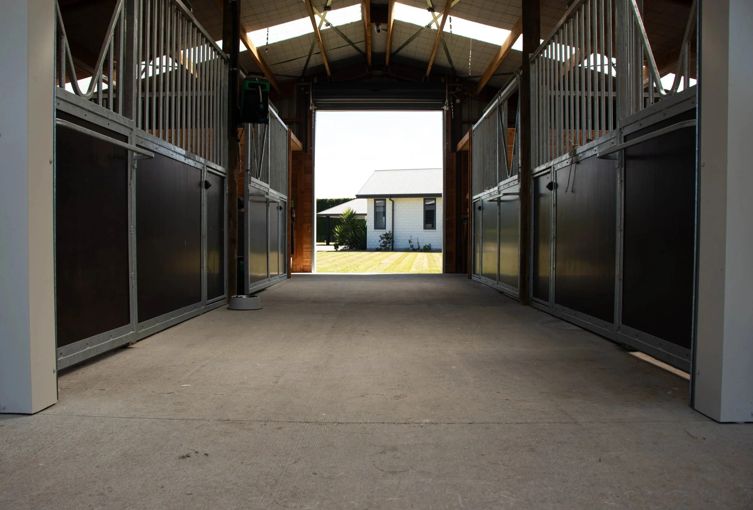 Inside horse stable with metal stall doors, view towards outside with a white house, trees, and a lawn in the background.