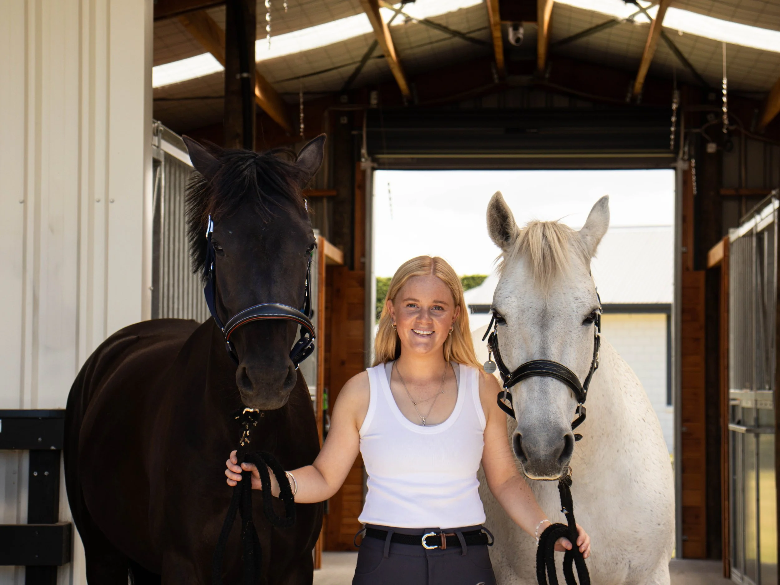 A woman smiling and holding two horses on leashes inside a barn, with one black and one white horse.
