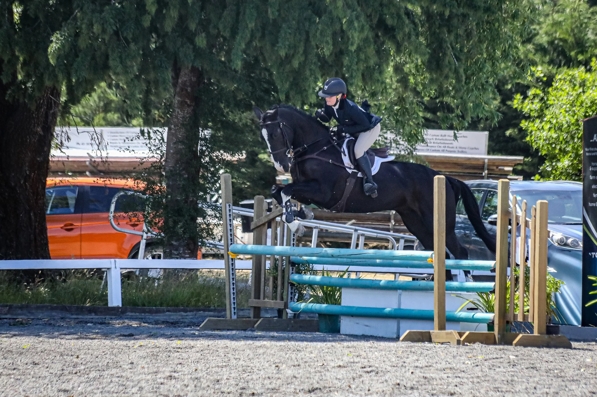 A rider in equestrian attire and a helmet jumping a black horse over a show jumping obstacle in an outdoor arena.