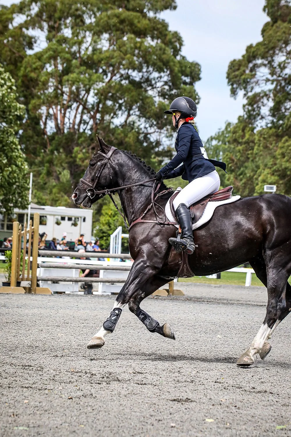 A female equestrian in formal riding attire riding a dark horse during a competition at an outdoor arena.