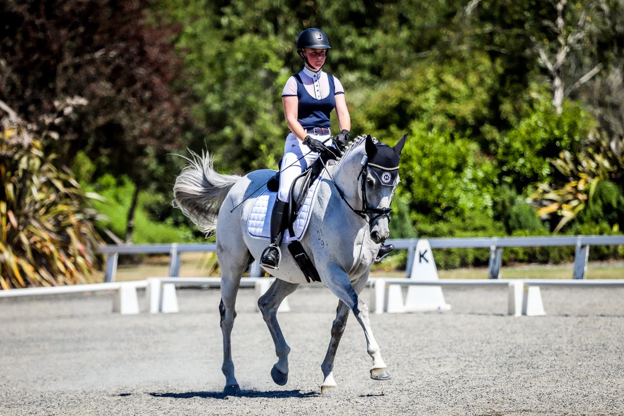 A female equestrian rider wearing a helmet, navy and white riding attire, and black gloves, riding a gray horse on a dressage arena with trees in the background.