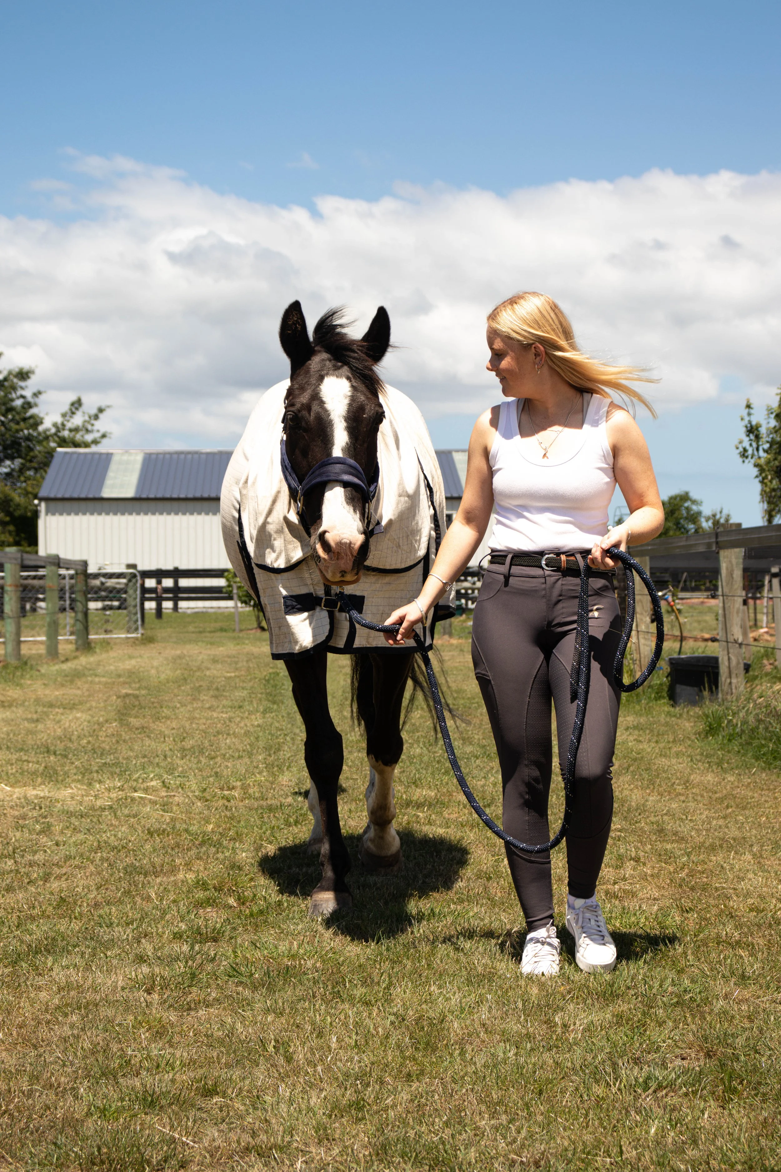 A woman walking a black and white horse on a grassy field, with a clear blue sky and a barn in the background.