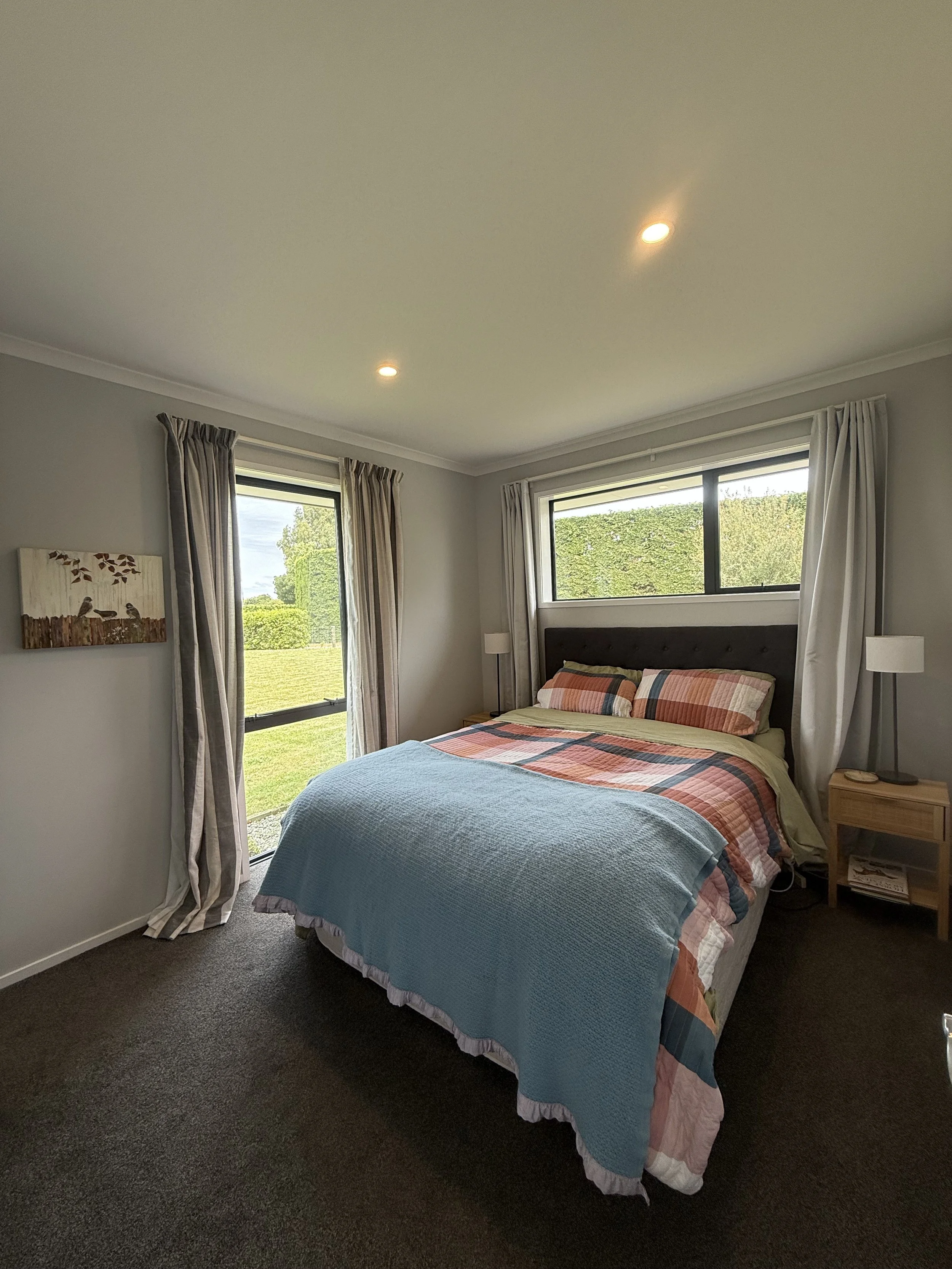 A bedroom with a bed covered in a colorful plaid quilt and pillows, two nightstands with lamps, two windows with curtains overlooking a green yard and hedge, and a painting on the wall.