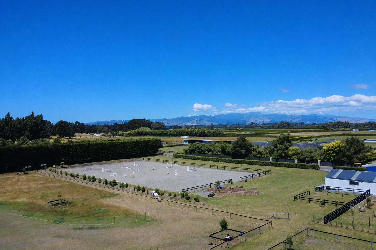 A landscape with a grid of outdoor horse jumping rings, a small white building, fenced paddocks, and a mountain range in the background under a clear blue sky.