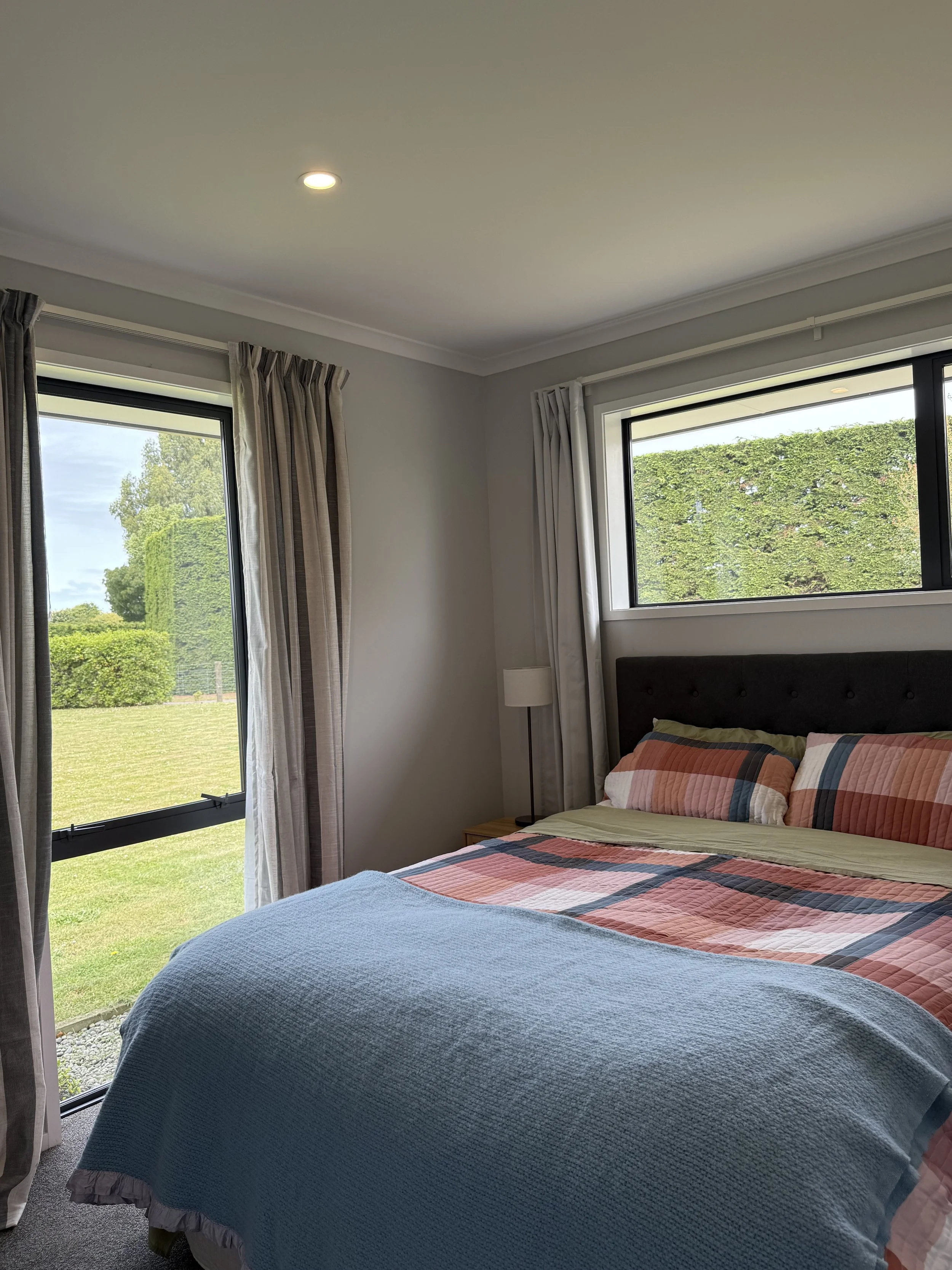 A bedroom with a bed covered in a plaid comforter and pillows, a small bedside table with a lamp, and large windows with curtains showing outdoor greenery.